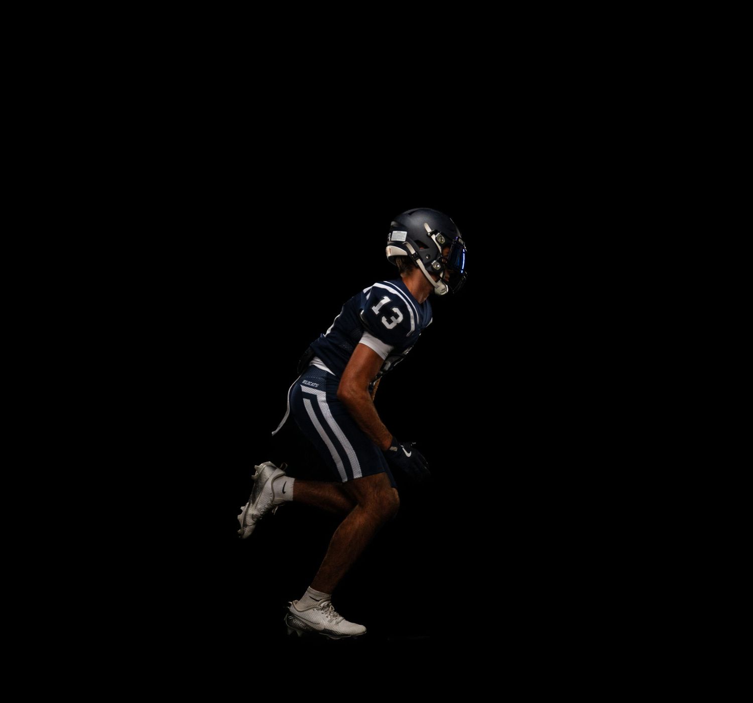 Football player in dark blue uniform mid-sprint against a black background.