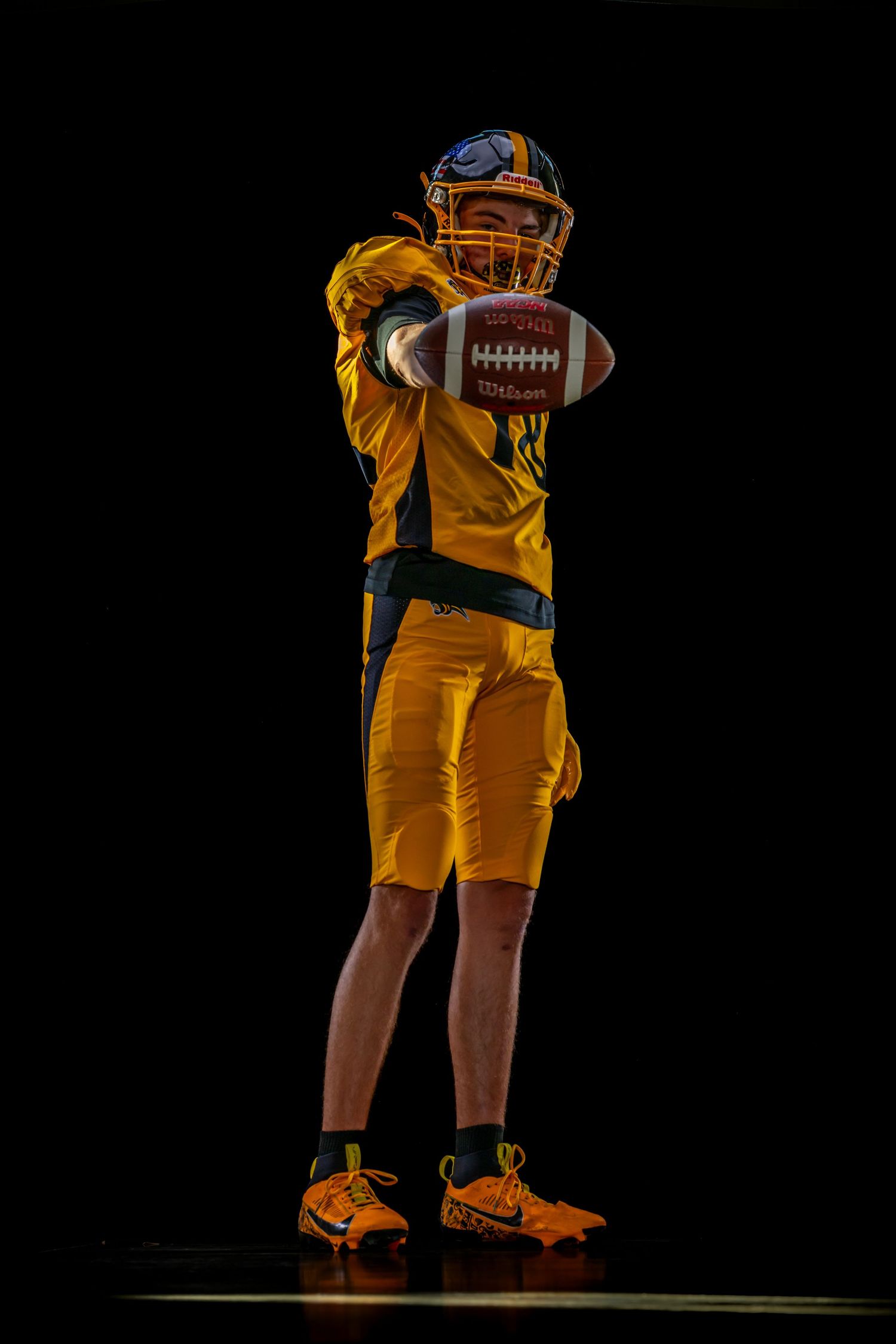 American football player in yellow uniform holds Wilson football toward camera against black background.