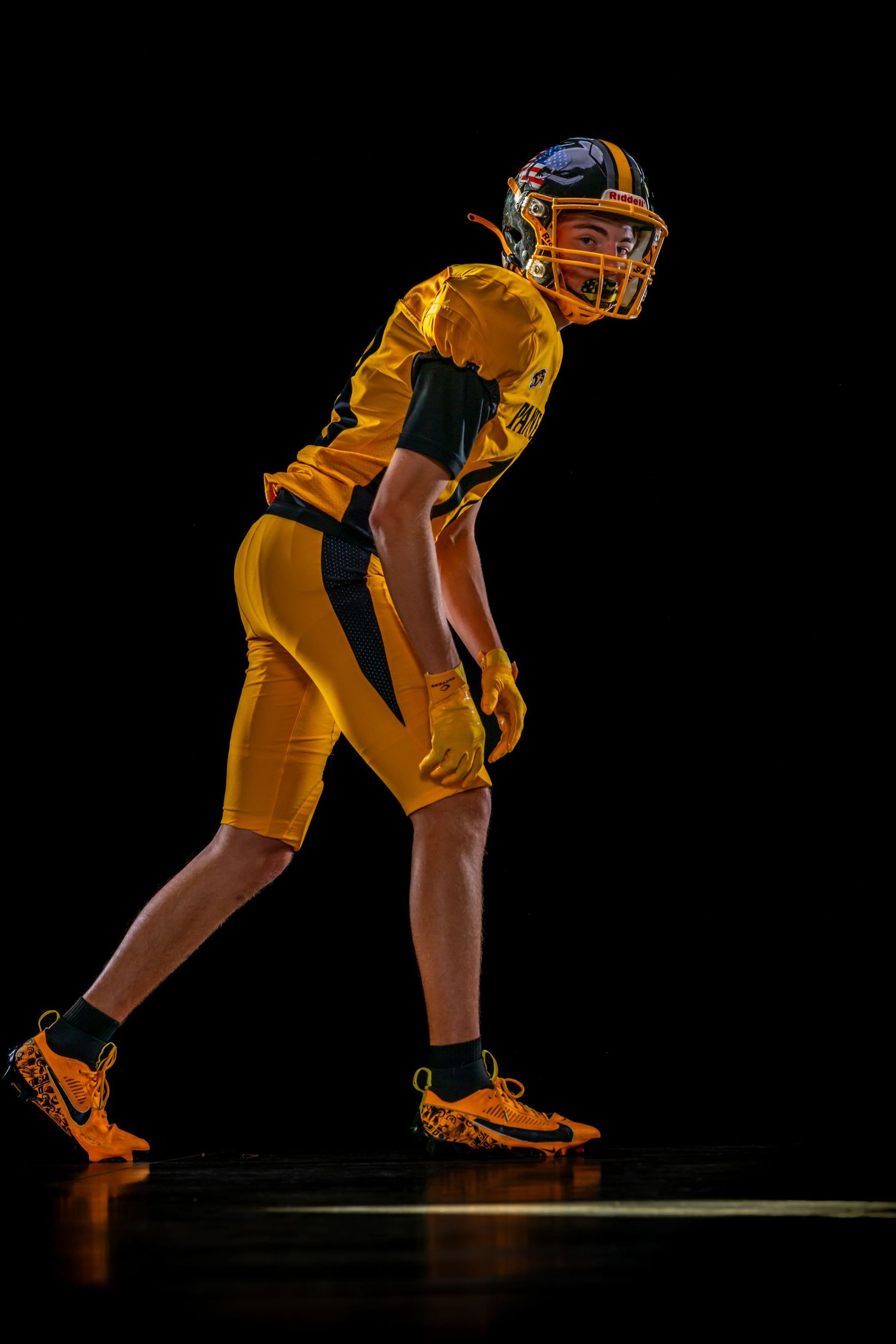 Youth football player in gold uniform and helmet in defensive stance on dark background.
