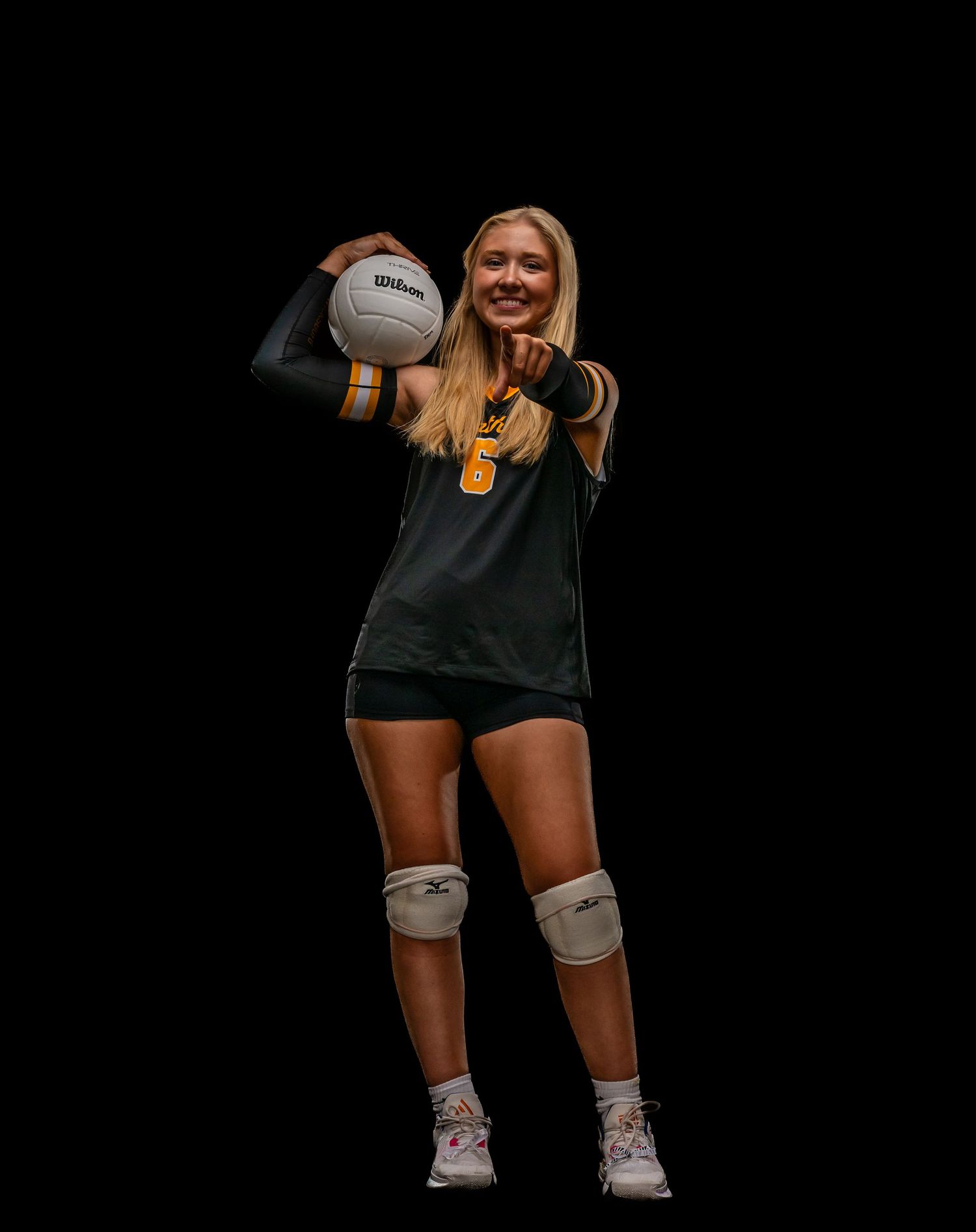 Blonde female volleyball player in black and gold uniform holding a Wilson volleyball, wearing knee pads.