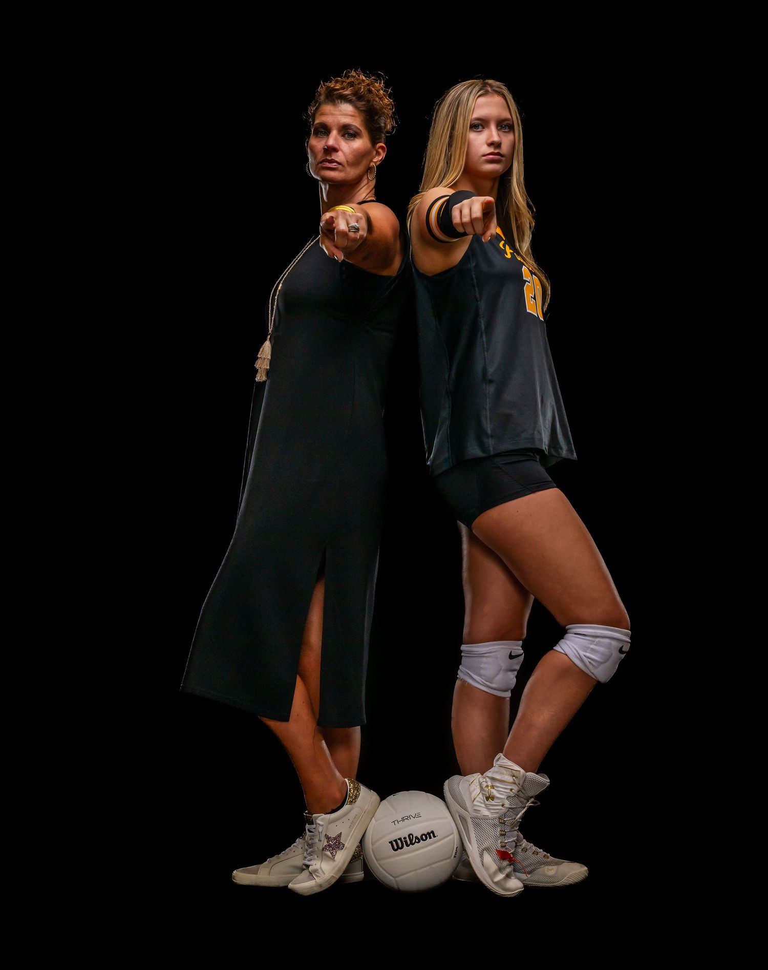 Two female volleyball players pose back-to-back pointing forward with Wilson volleyballs at their feet on black background.
