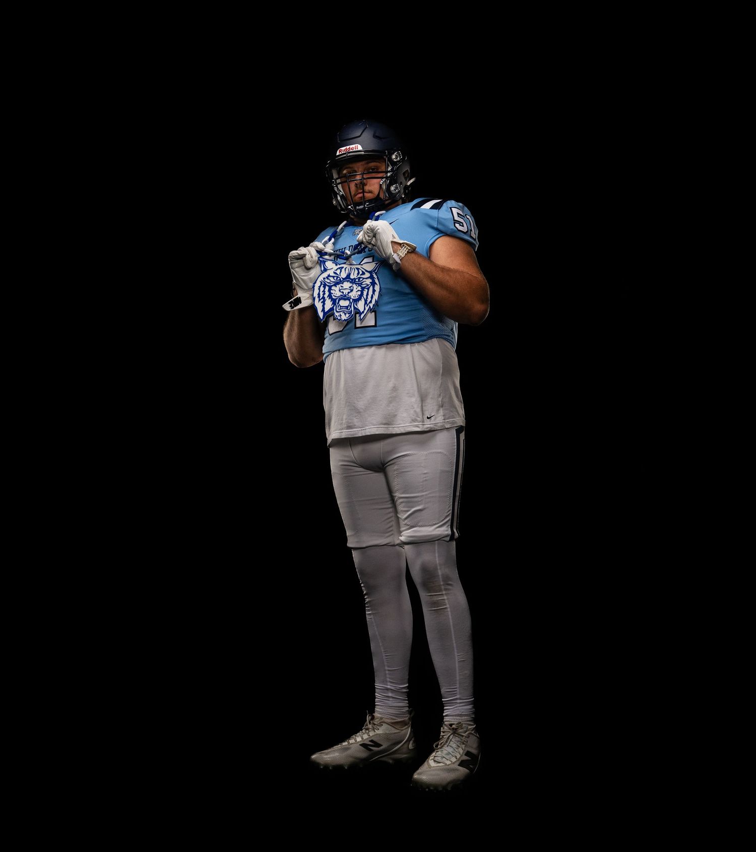 Football player in light blue jersey and gray pants poses in full uniform with helmet against black background.