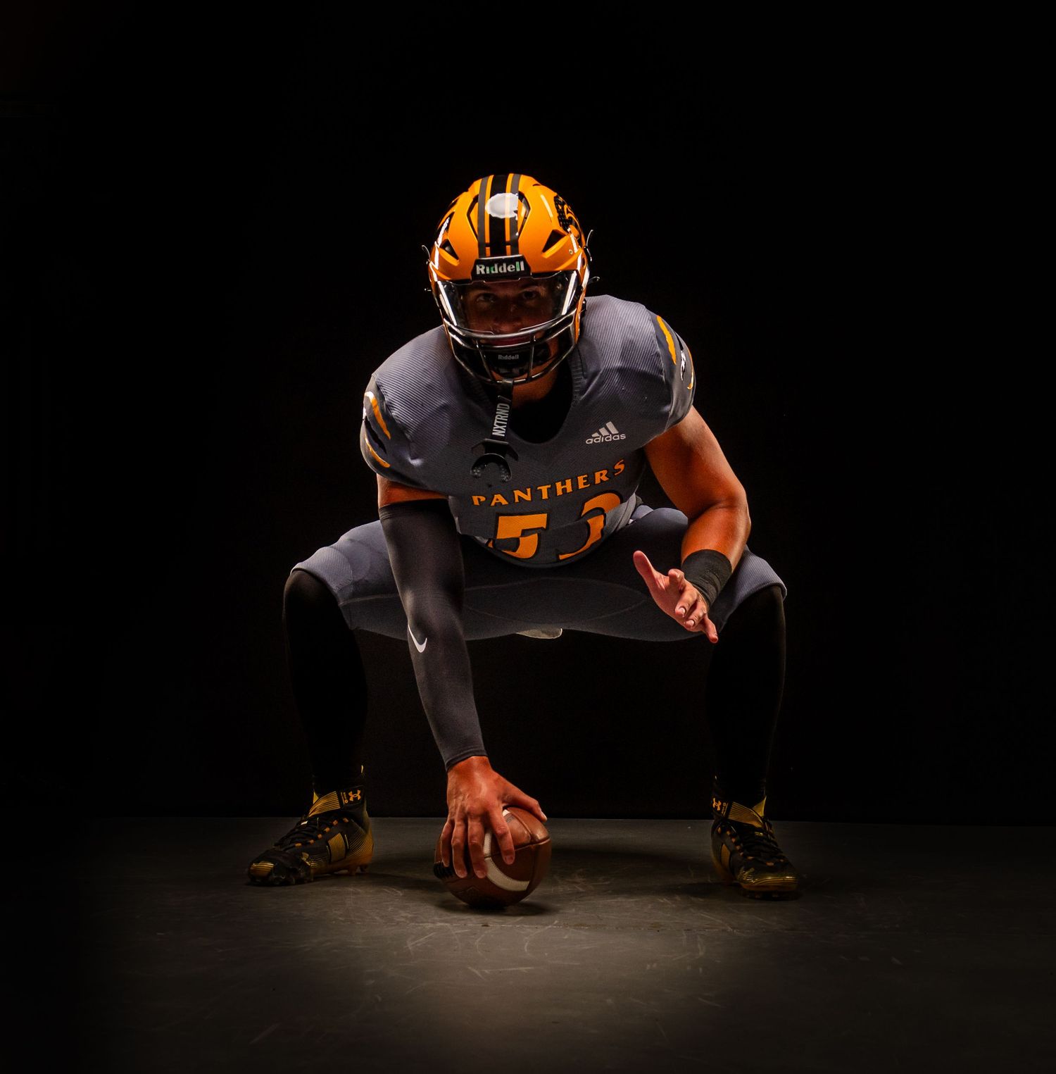 Football player in orange and black uniform crouches in dramatic lighting against dark background.