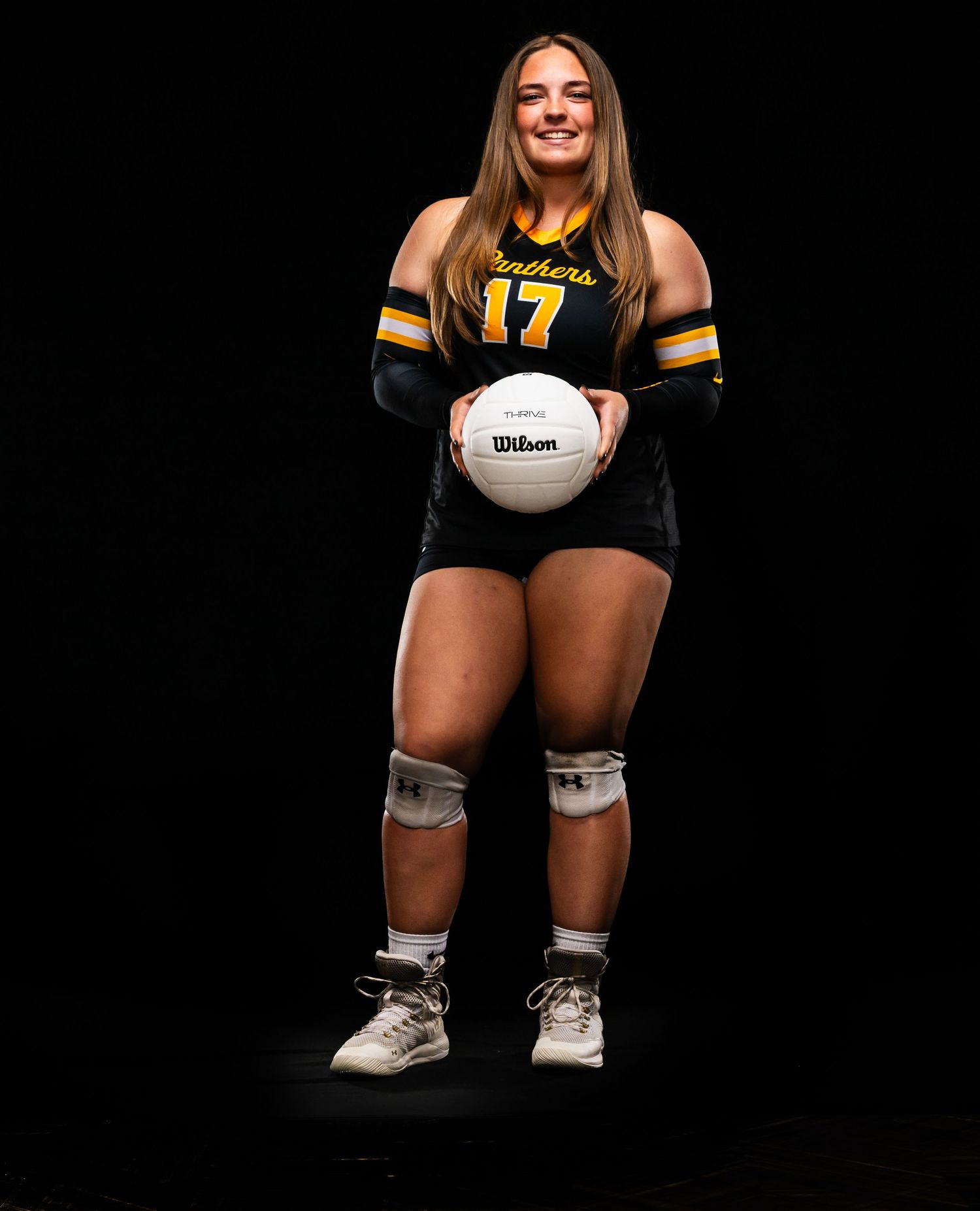 Volleyball player in black uniform crouches with ball against dark background.