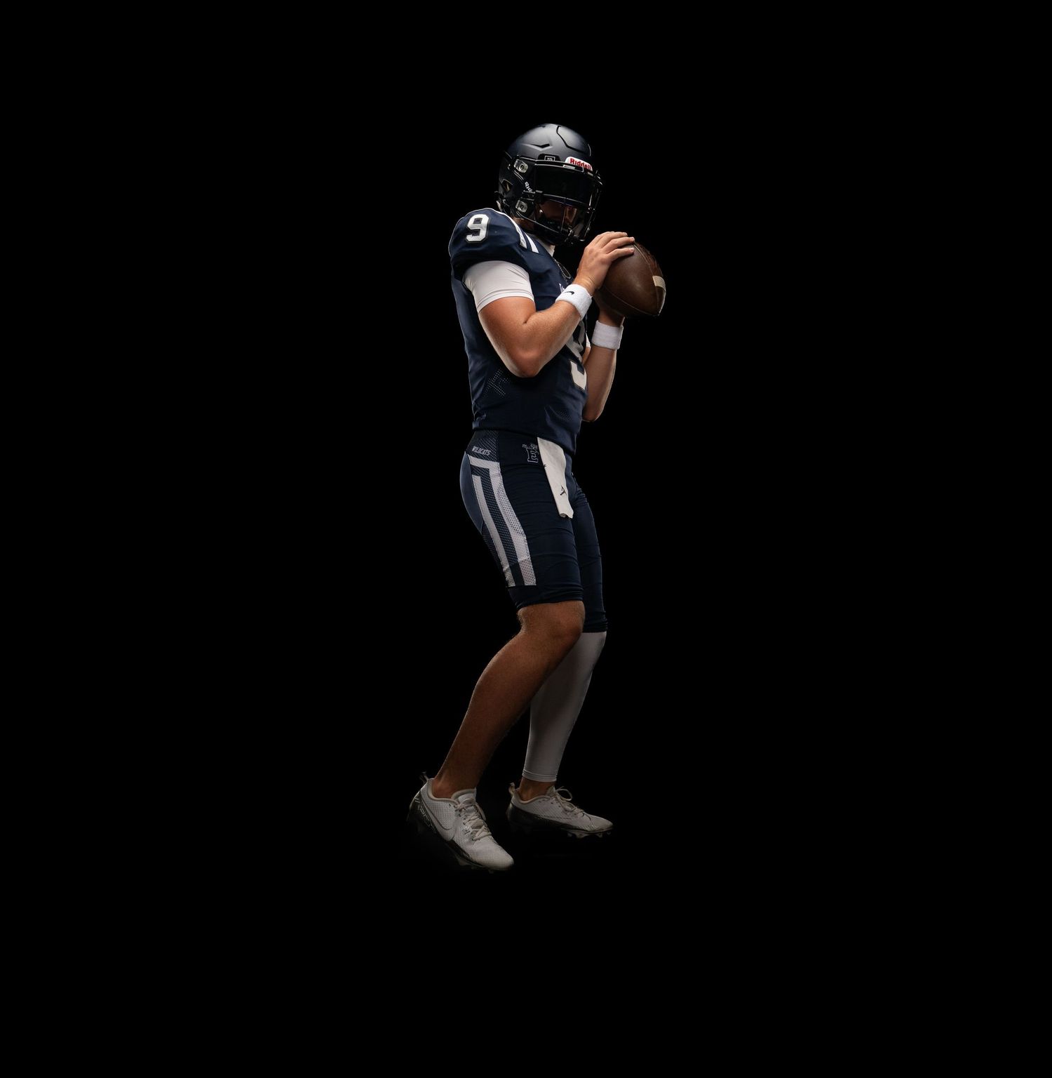 Football player in navy uniform holding ball in throwing stance against black background.