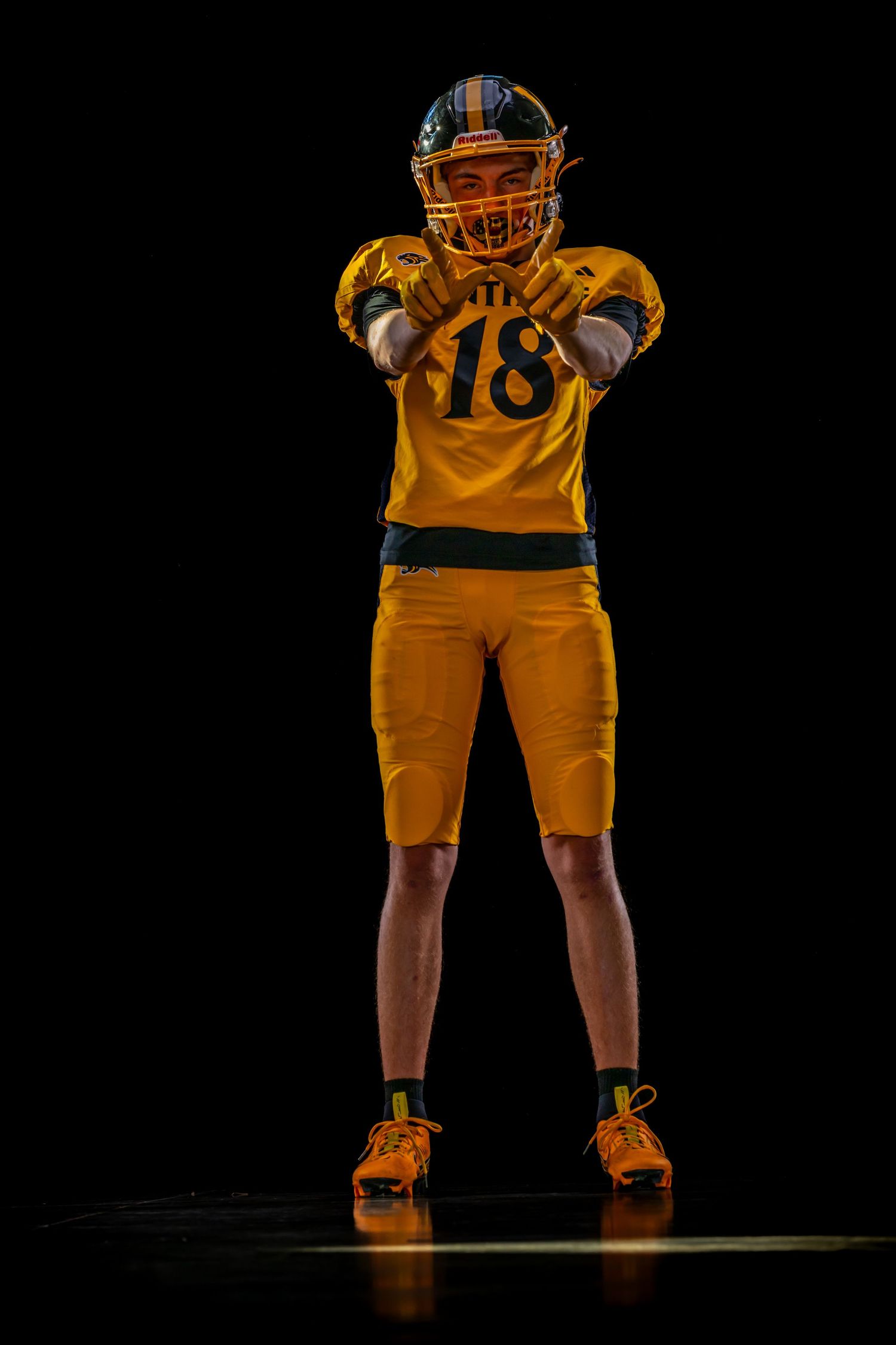 Football player wearing gold jersey number 18 poses with arms extended on dark background.