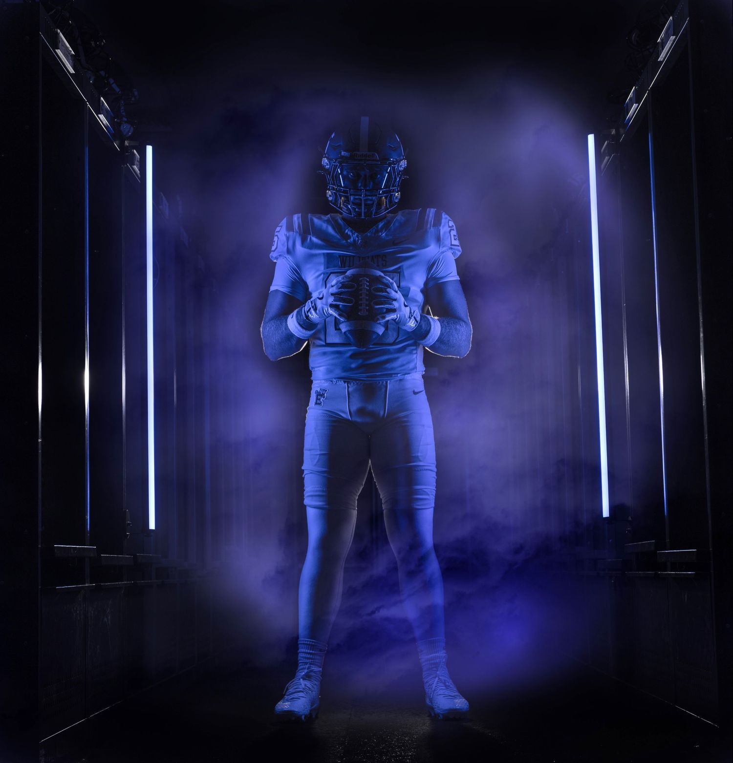 Football player in blue uniform and helmet stands dramatically in a dark tunnel with glowing neon lights and blue smoke.