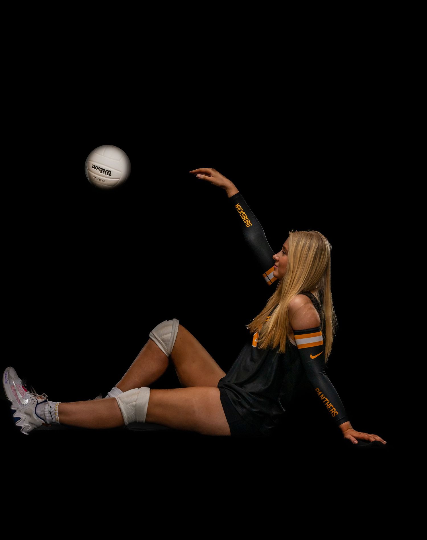 Female volleyball player in black Nike uniform diving to hit a white volleyball against a dark background.