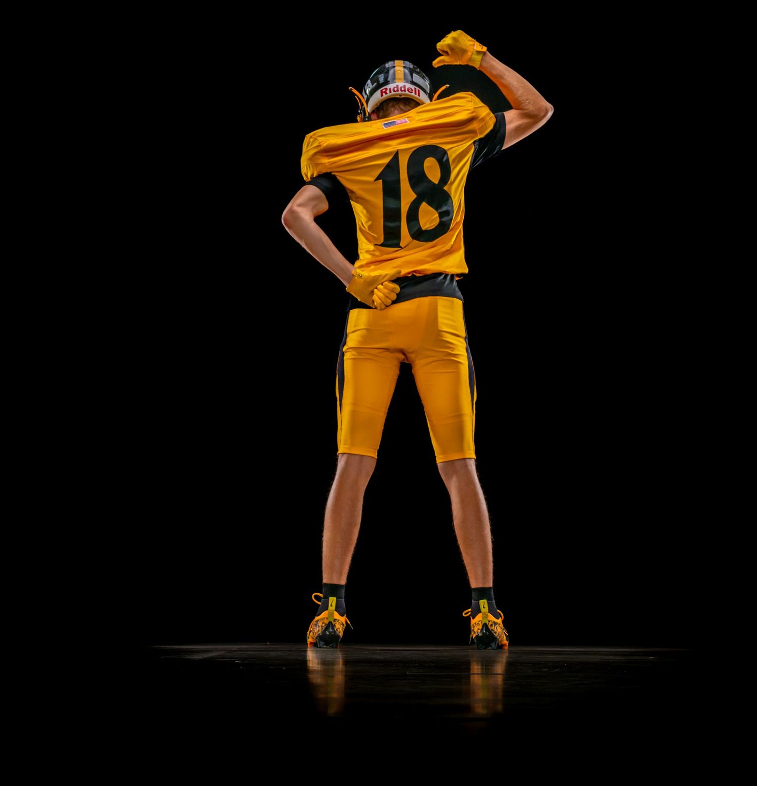 Football player #18 in gold uniform poses confidently from behind against black background.