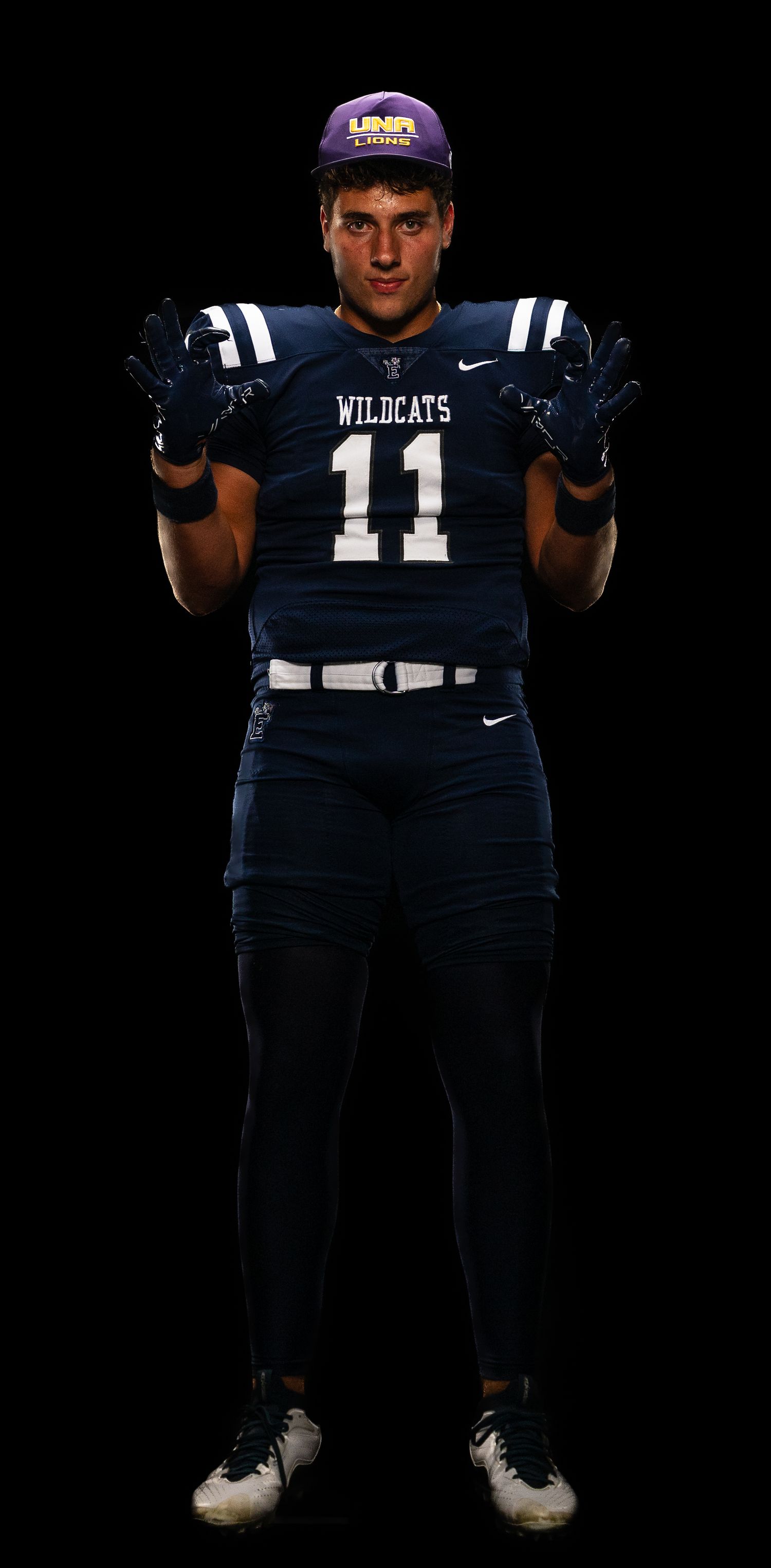 Football player wearing Wildcats number 11 navy jersey poses with UNR cap against black background.