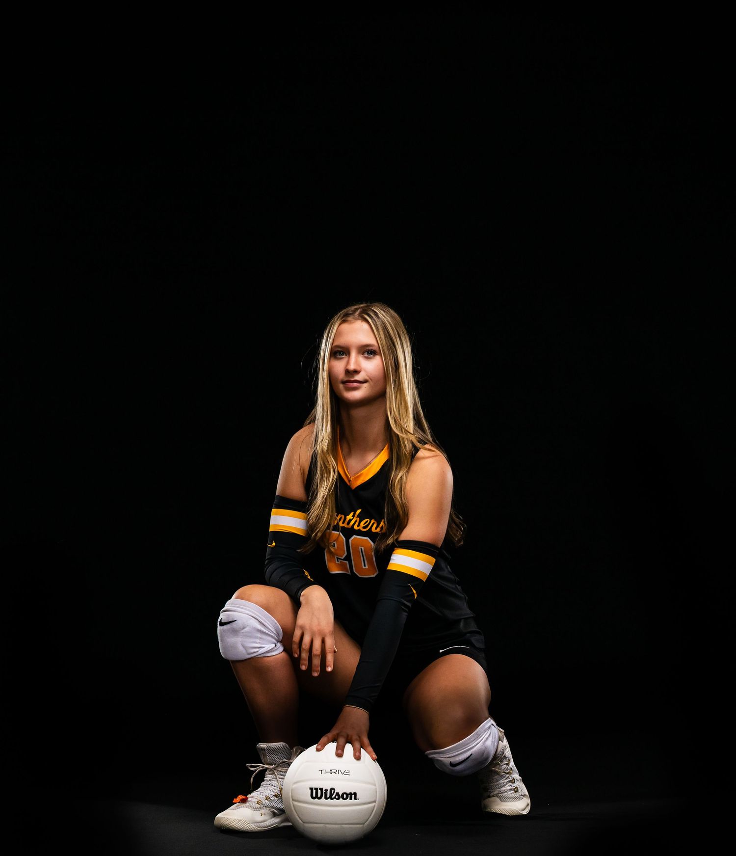 Female volleyball player in black and gold uniform crouching with Wilson volleyball on dark studio background.