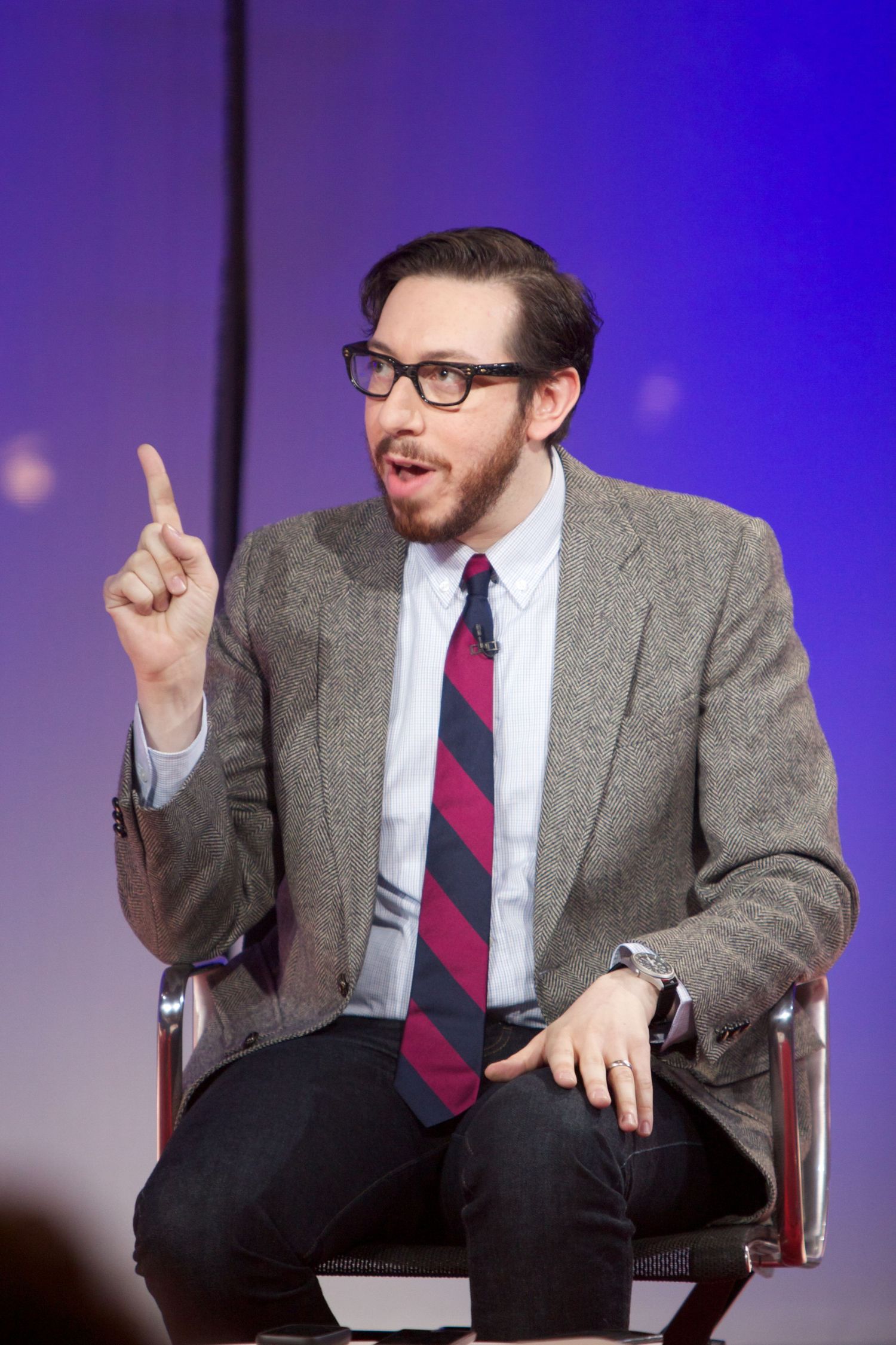 Speaker gesturing while seated in chair during presentation with blue stage lighting.