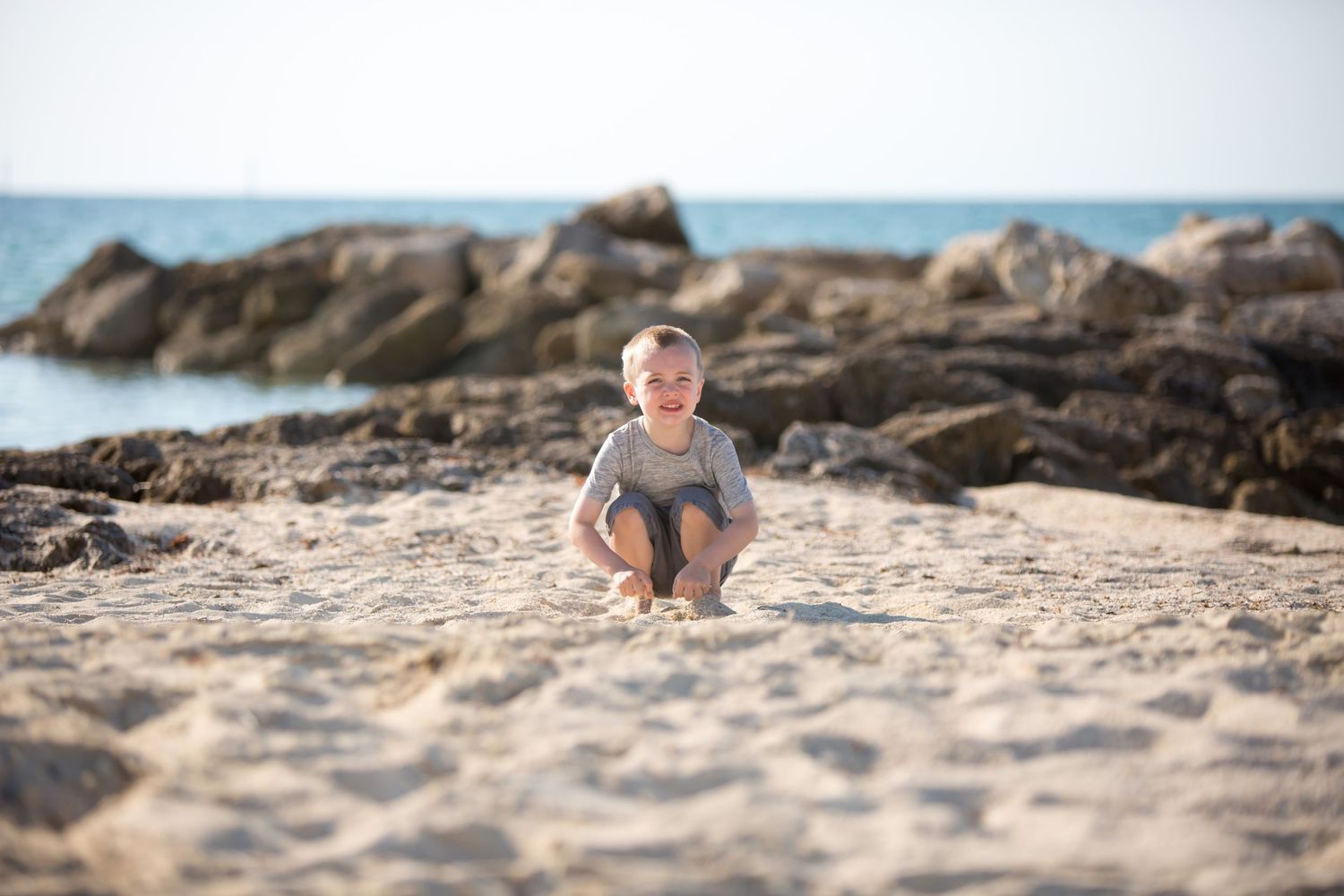 Small figure sitting on beach sand with rocky coastline in background.