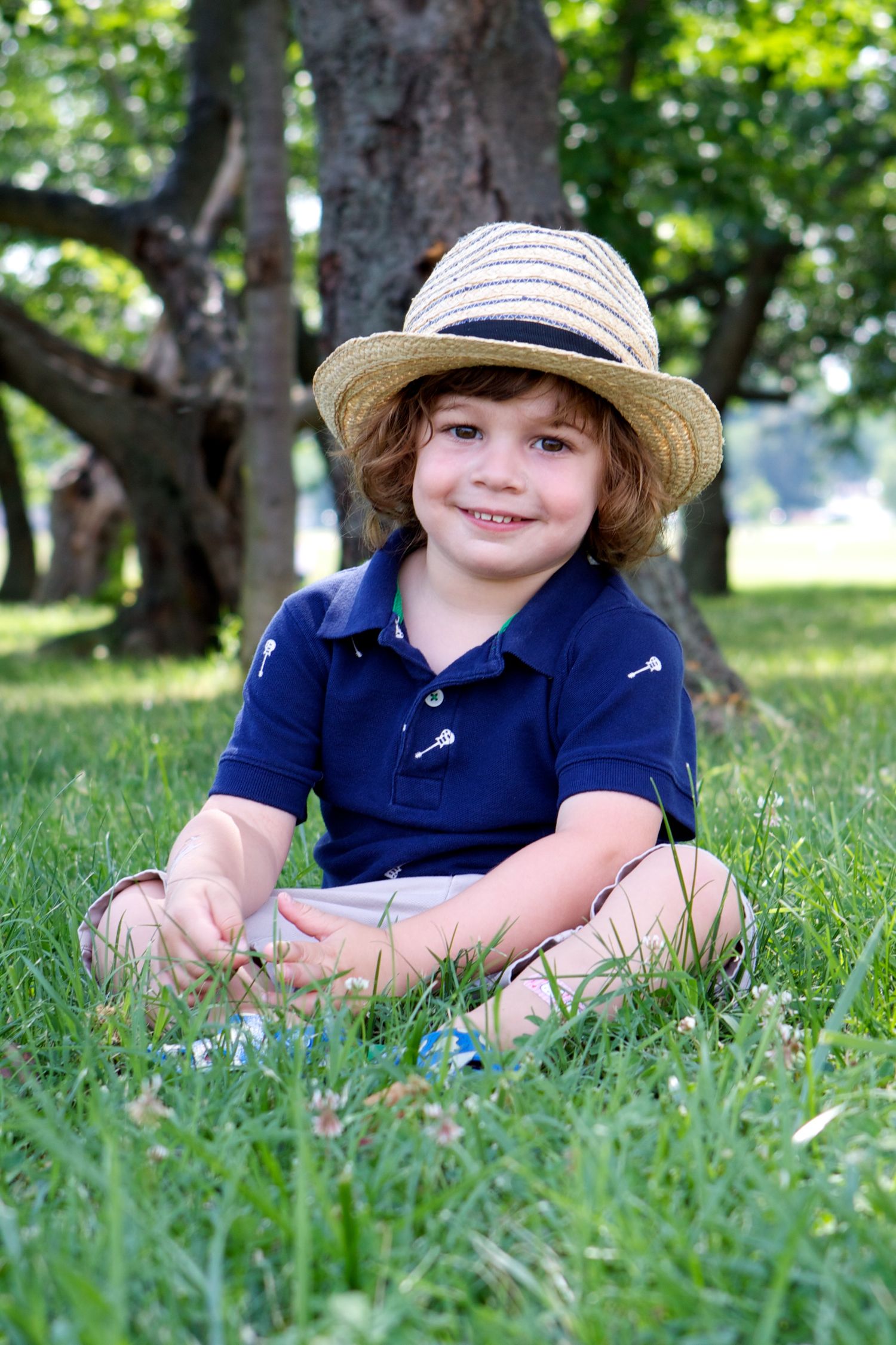 Child wearing straw hat and navy polo shirt sitting in grass under trees.