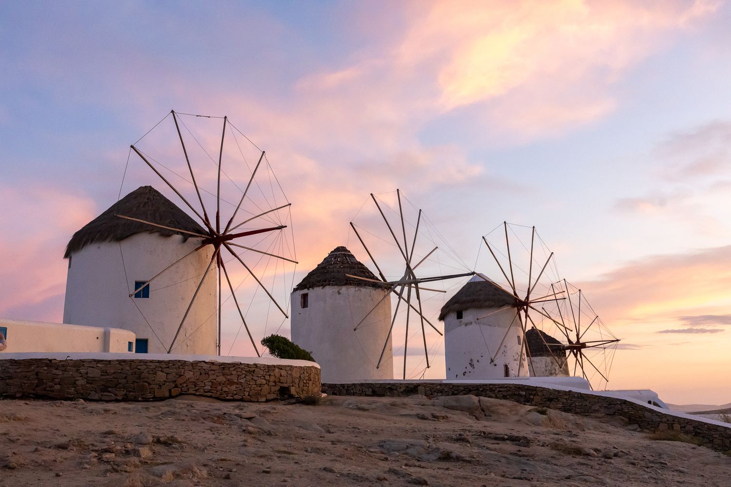 Traditional white windmills line the coast of Mykonos at sunset with pink and orange sky.