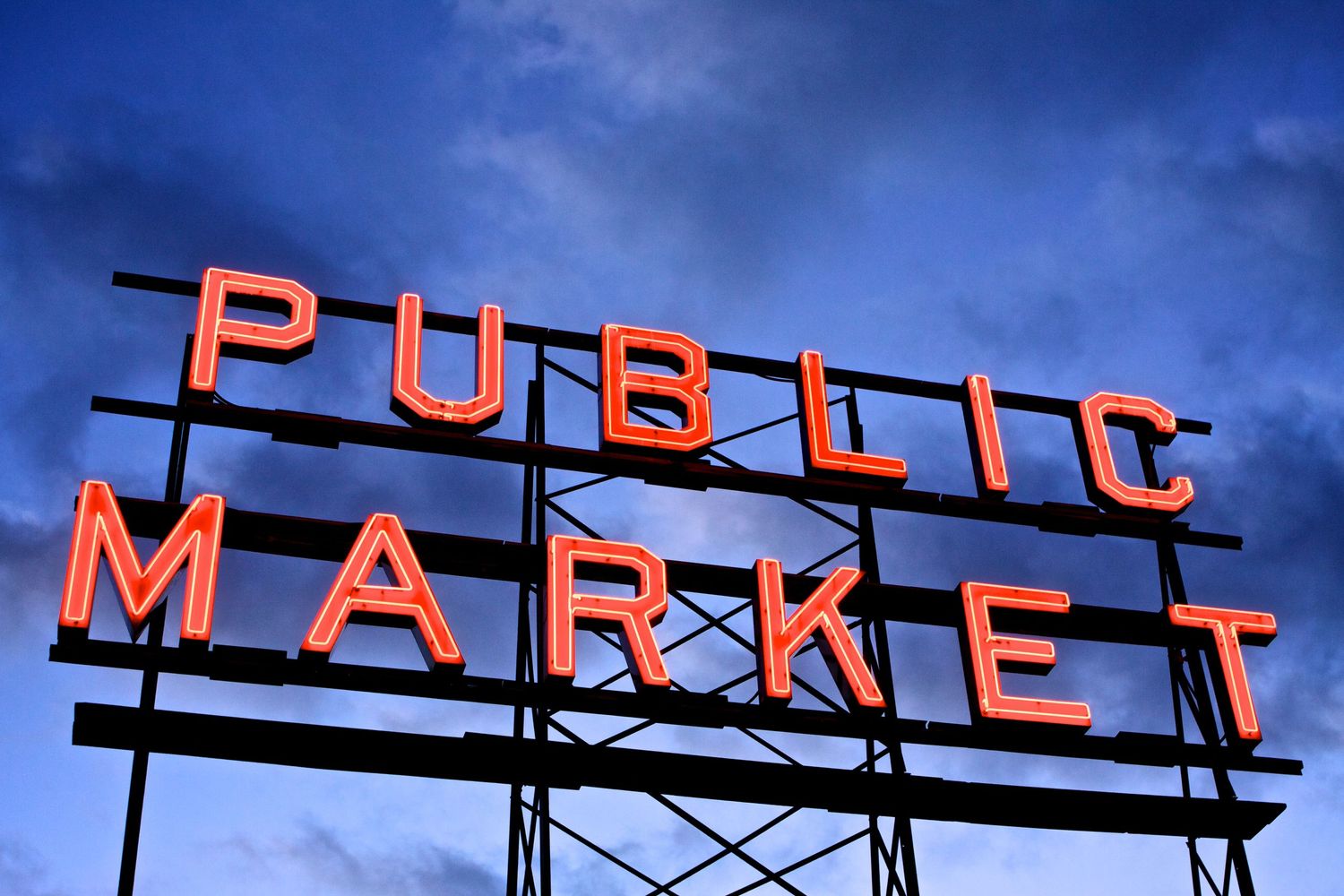 Illuminated red neon sign spelling PUBLIC MARKET against a dark blue evening sky.