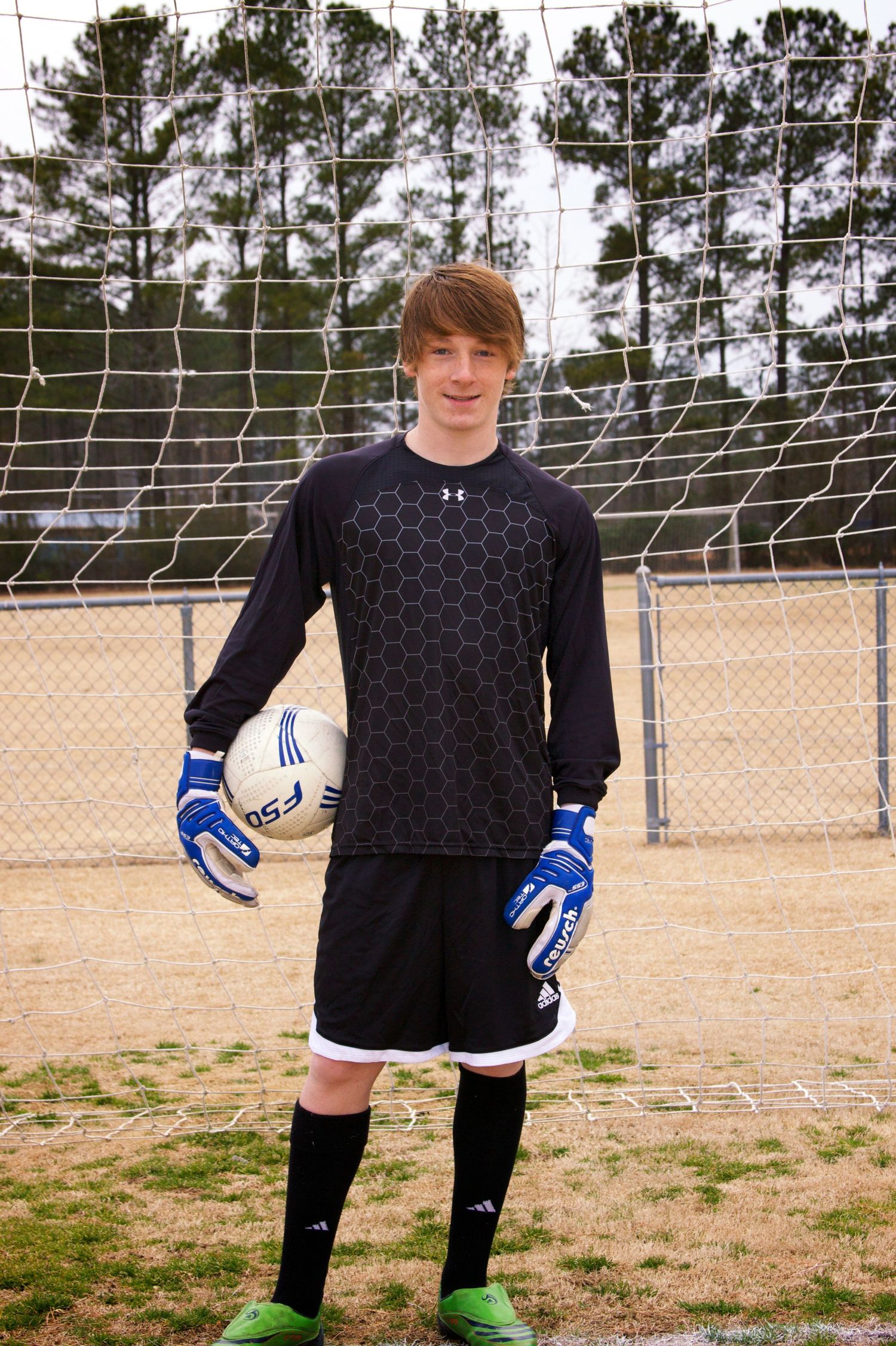 Young goalkeeper in training gear poses with soccer ball on field with goal net behind.