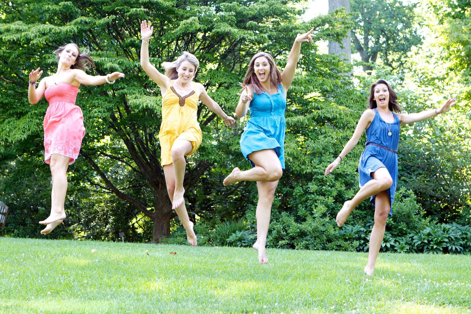 Four people in bright dresses jumping simultaneously in garden setting.