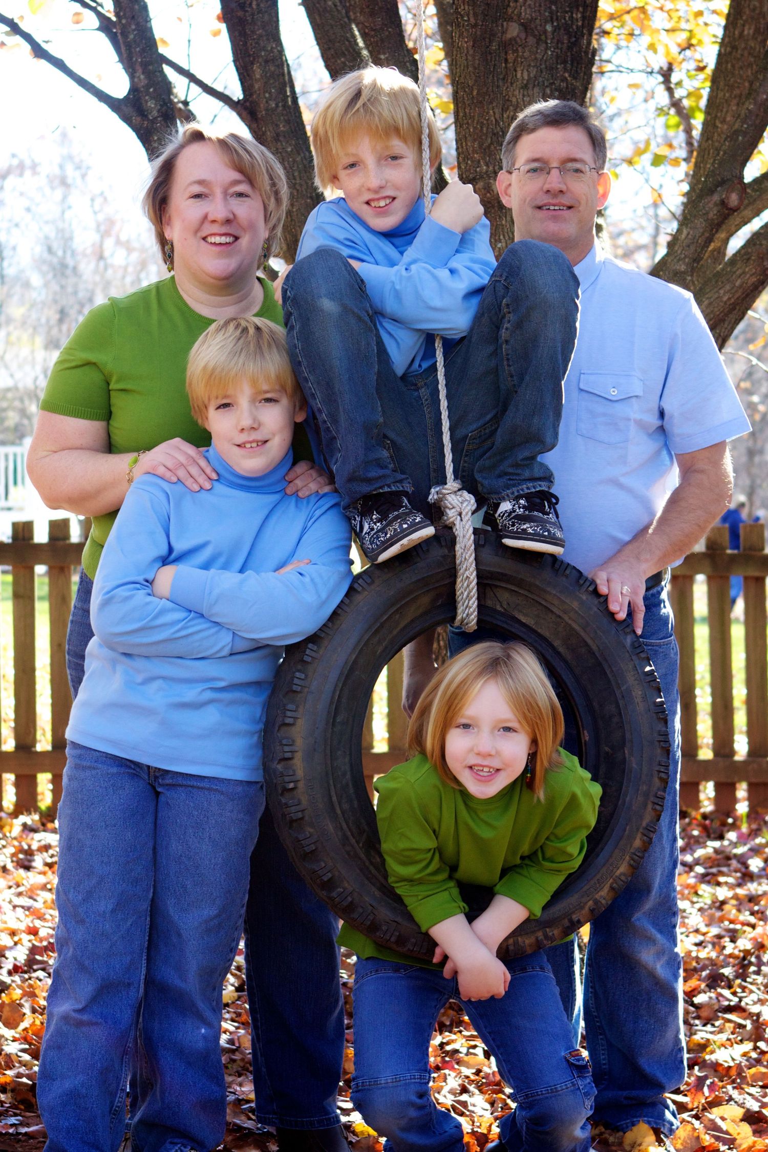 Family group poses together around tire swing in autumn setting with fallen leaves.