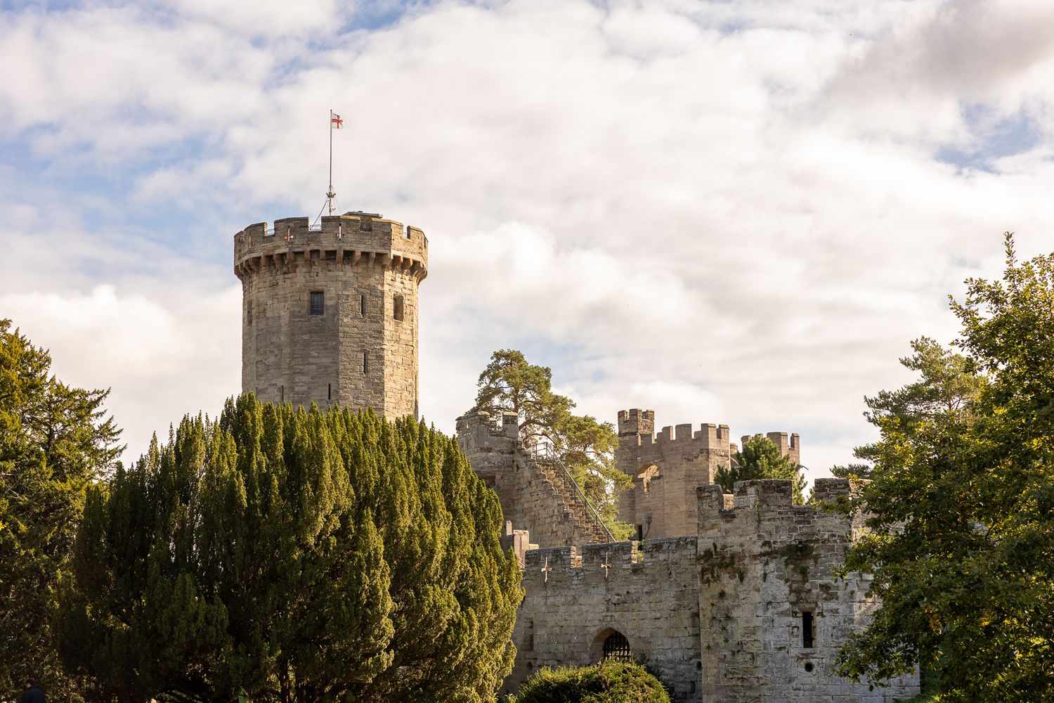 Medieval stone castle tower and walls surrounded by green trees under cloudy sky.