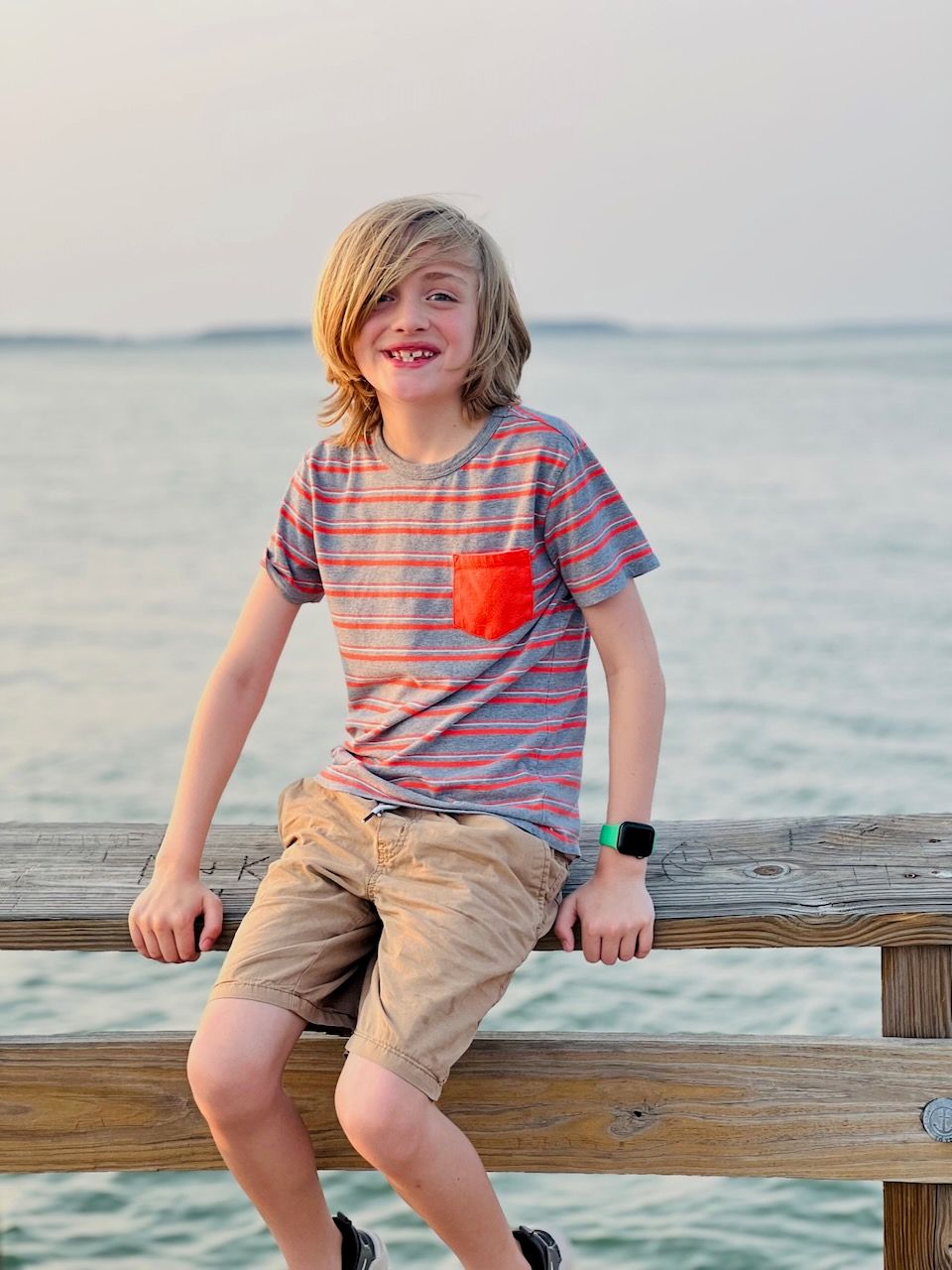 Child in striped shirt sitting on wooden pier overlooking calm ocean at sunset.