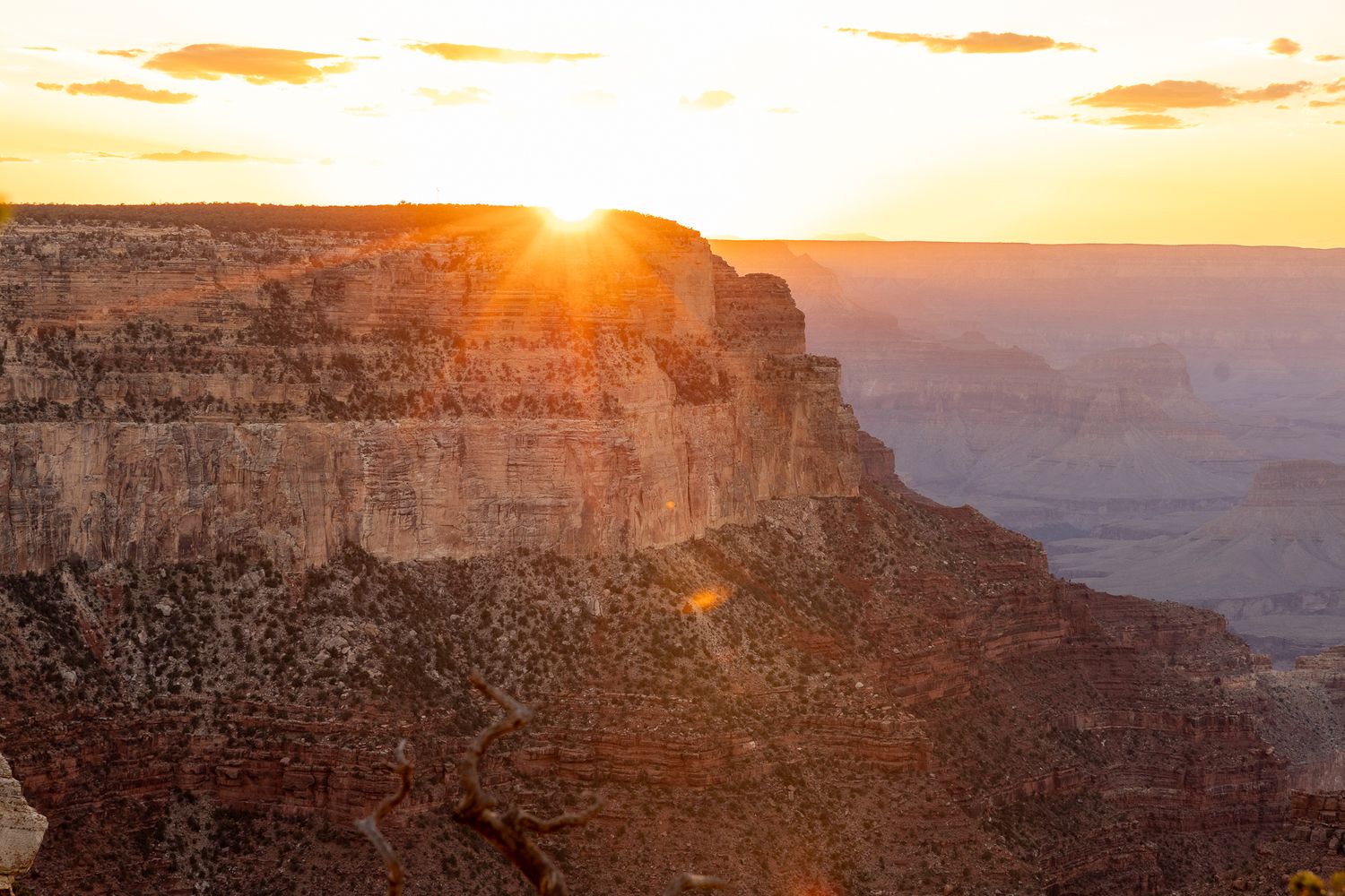 Dramatic sunset over the Grand Canyon with sunburst peeking through layered rock formations.