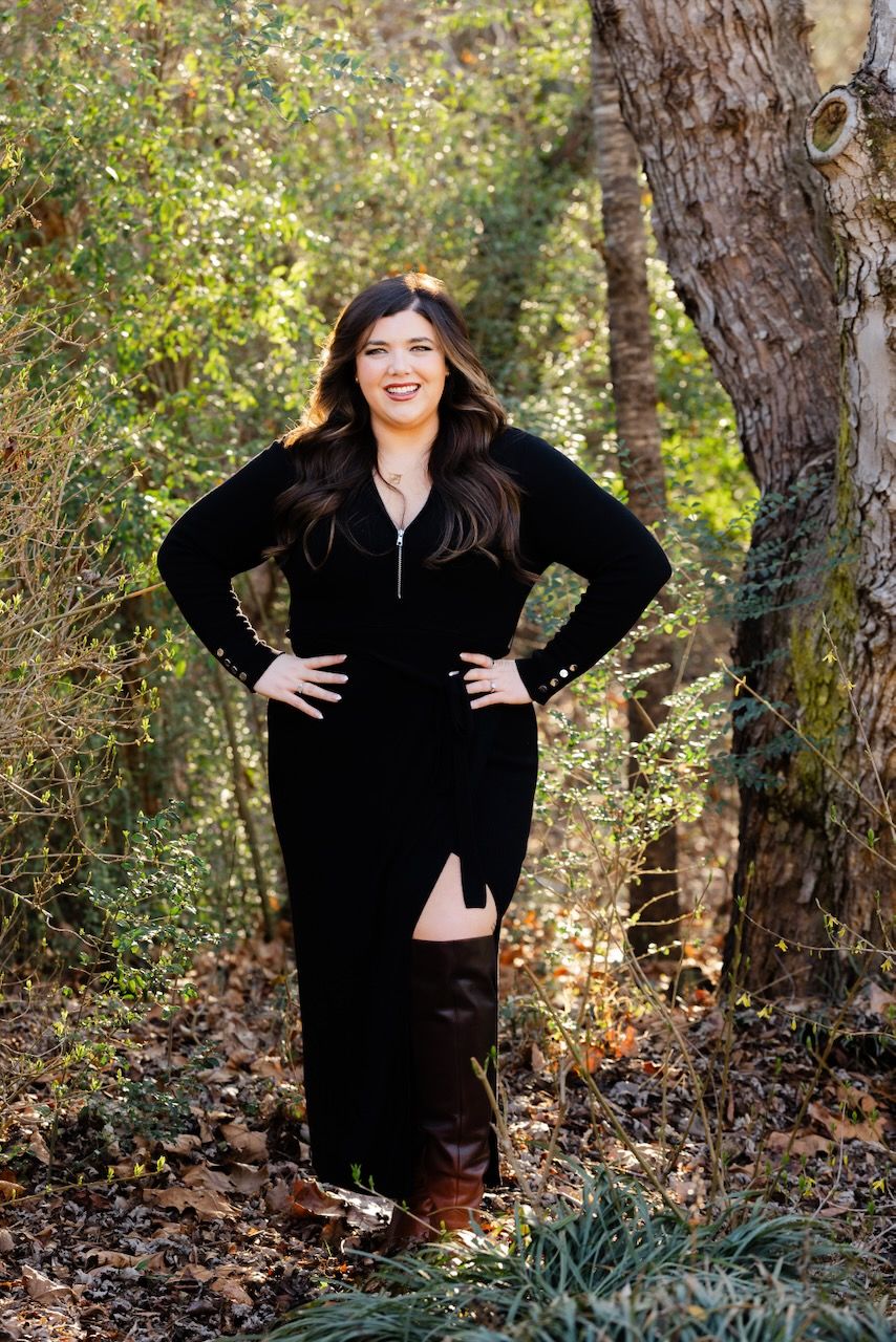 A person in a black velvet dress poses confidently against a natural wooded backdrop with autumn foliage.