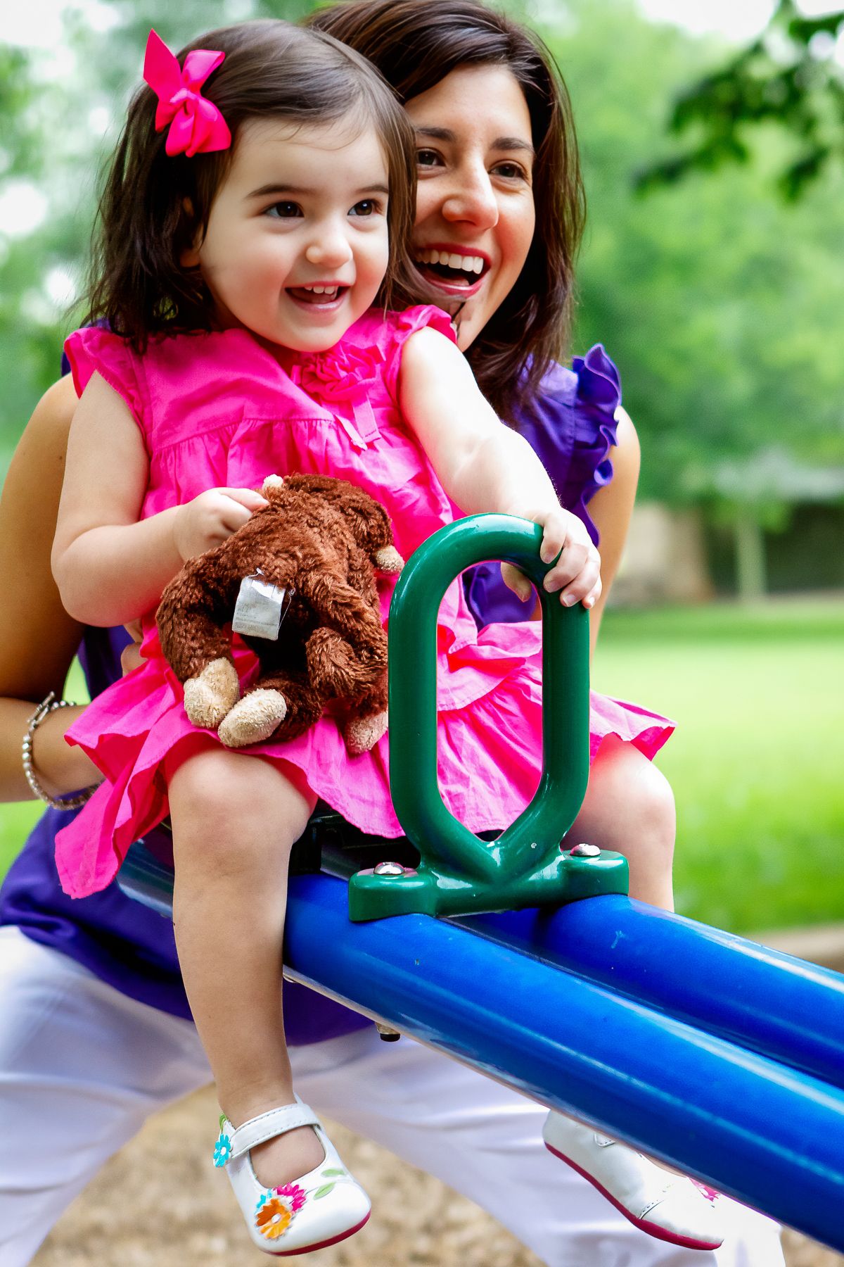 Mother and toddler girl in pink dress holding teddy bear smiling on playground seesaw in green park.