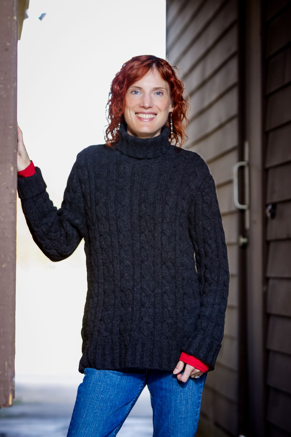 Smiling redhead woman in black cable-knit turtleneck sweater and jeans leaning against a building exterior.