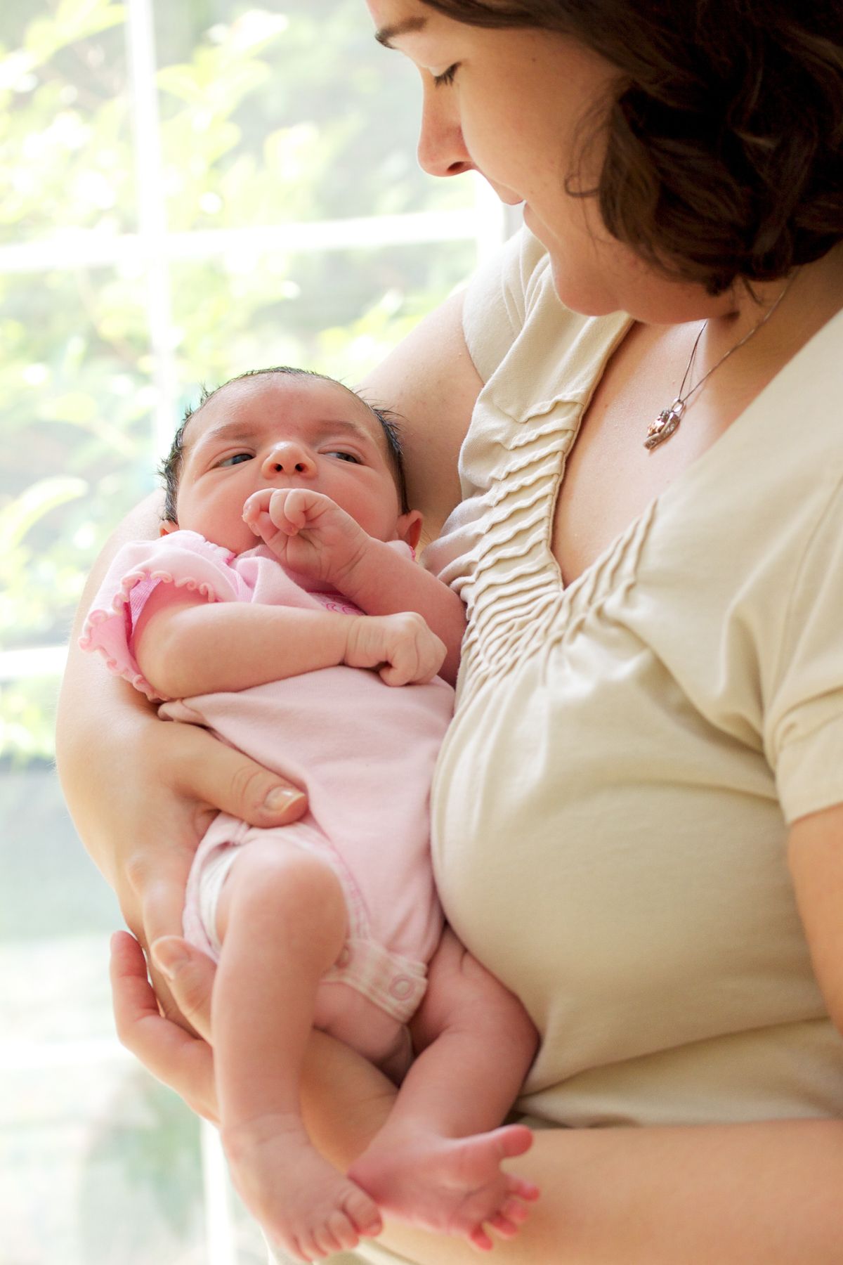 A mother tenderly holds her newborn baby dressed in pink, gazing lovingly at the infant near a bright window.