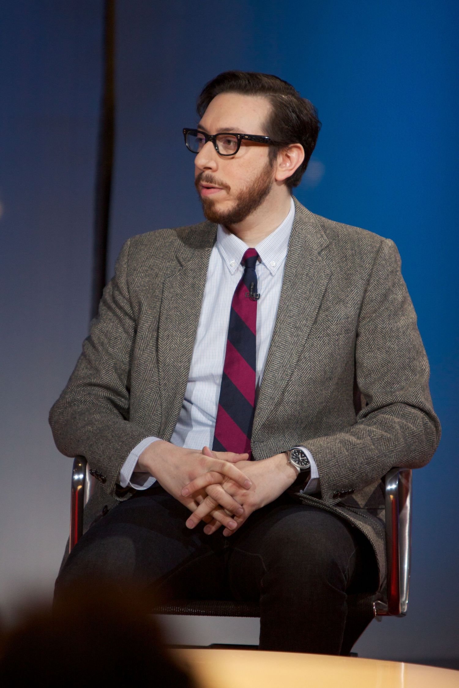 Professional seated portraits in a tweed suit and glasses against a blue background.