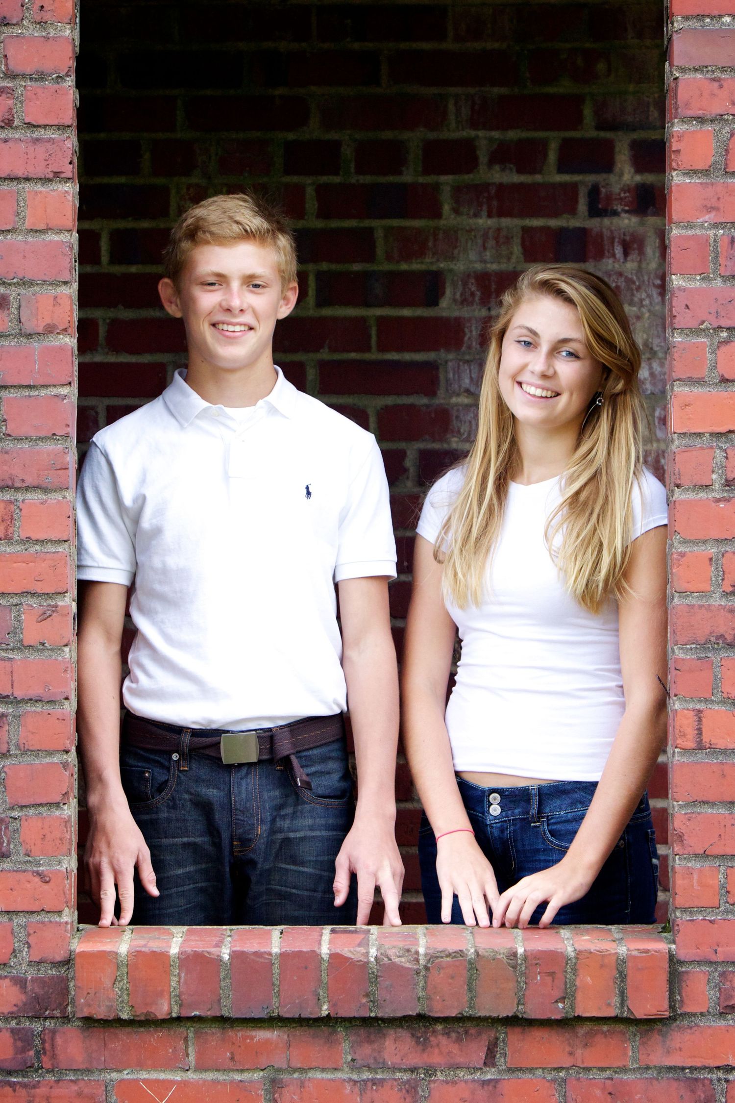 Two young people posing together in a brick window frame wearing casual white tops.