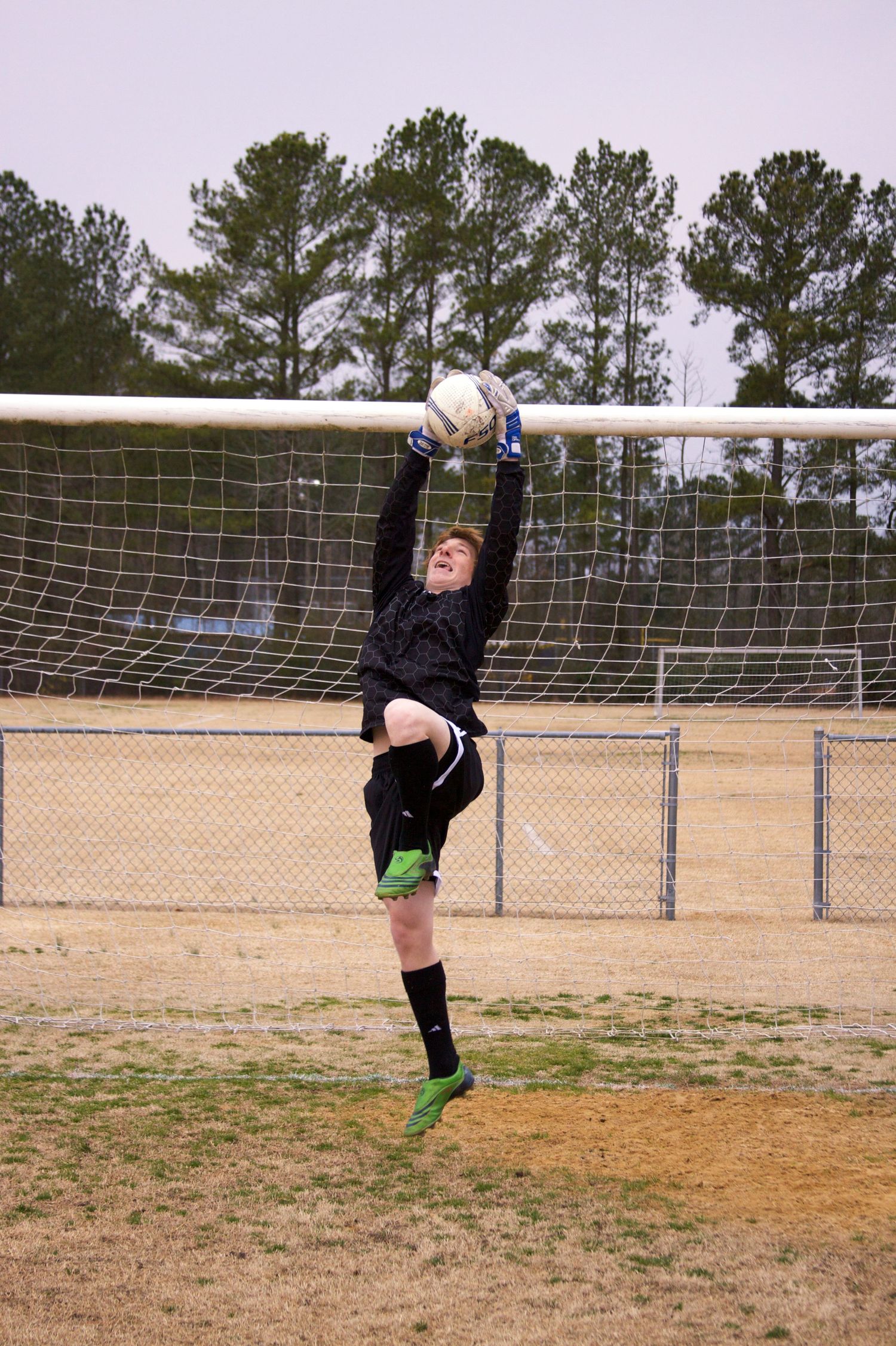 Soccer goalkeeper in black uniform practices diving saves on outdoor field with goal net.