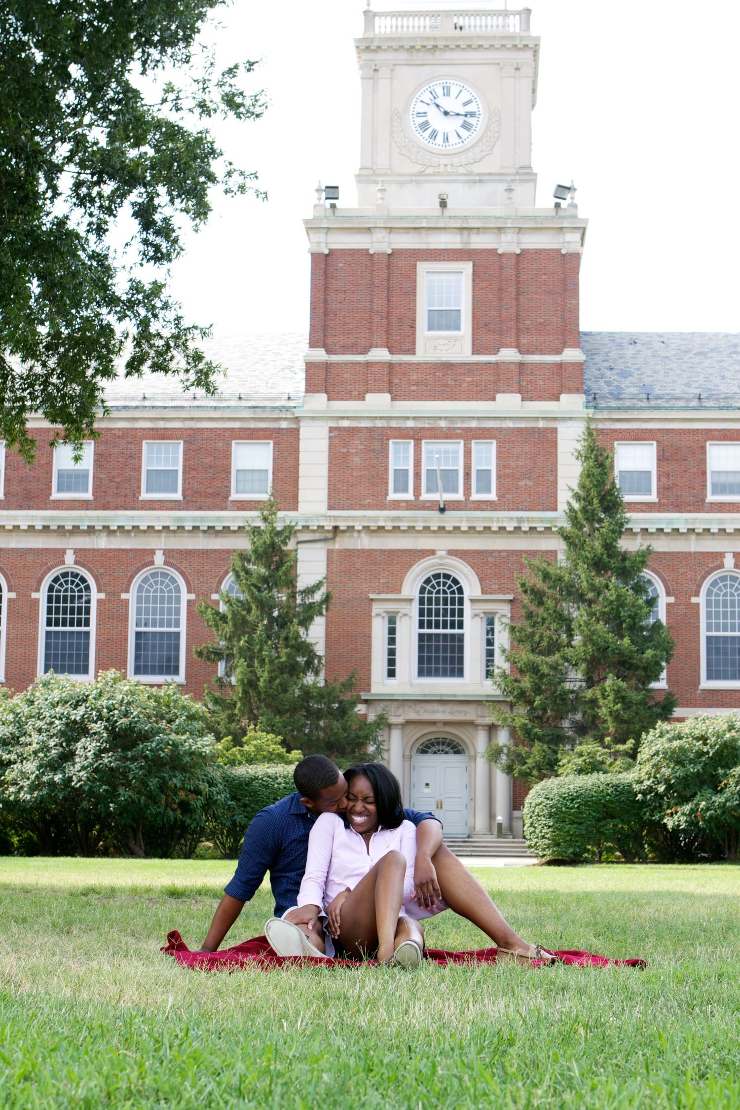 A couple sits on a blanket in front of a grand brick clock tower building.