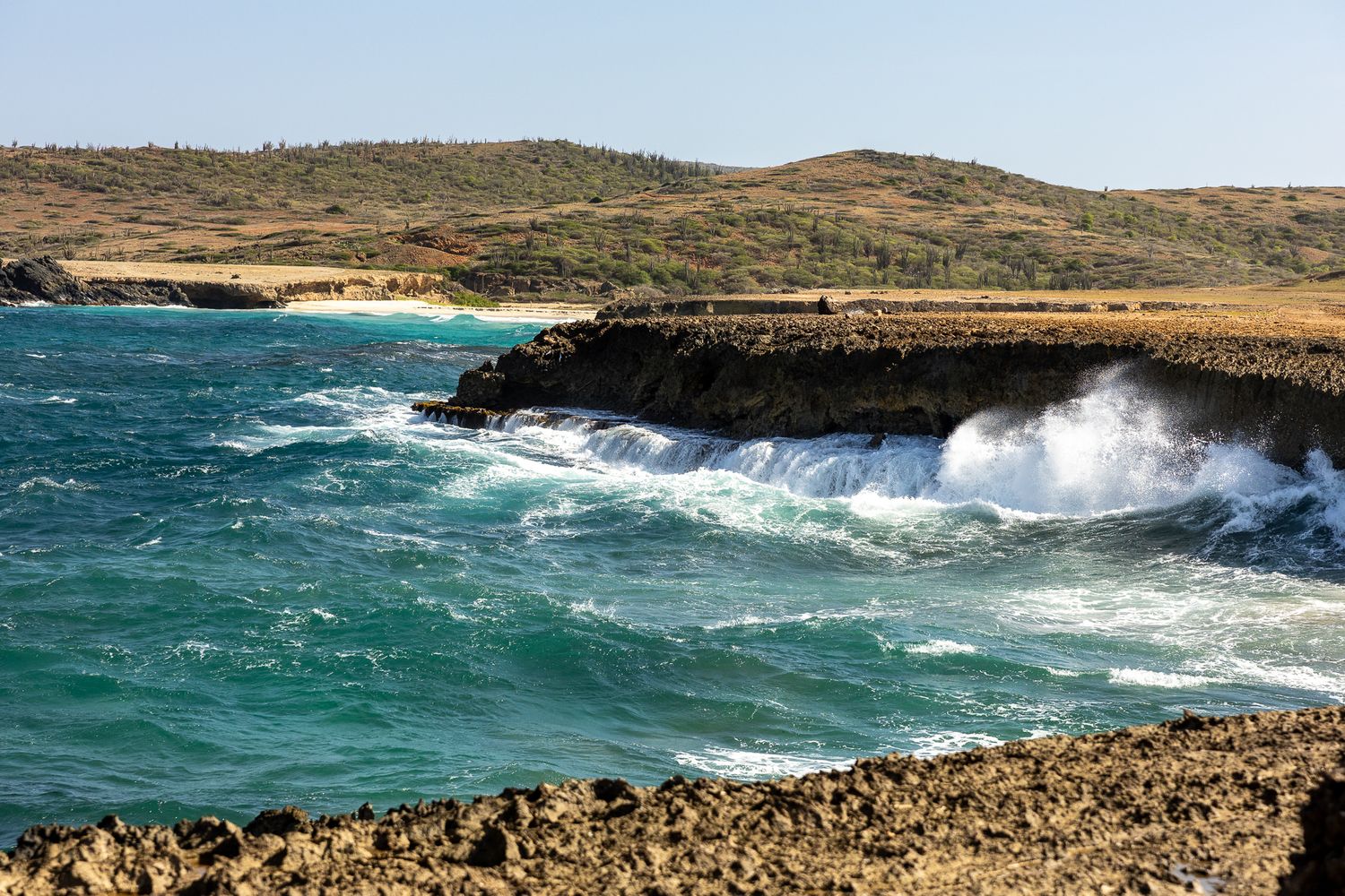 Rocky coastline with turquoise waves crashing against cliffs under bright sunlight.