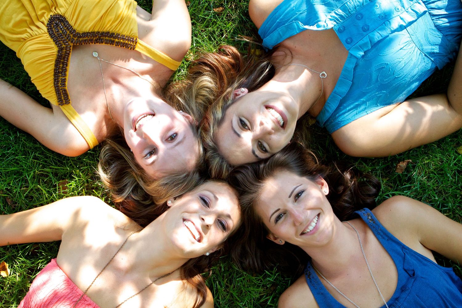 Four people lying on grass in circle formation wearing colorful summer outfits.