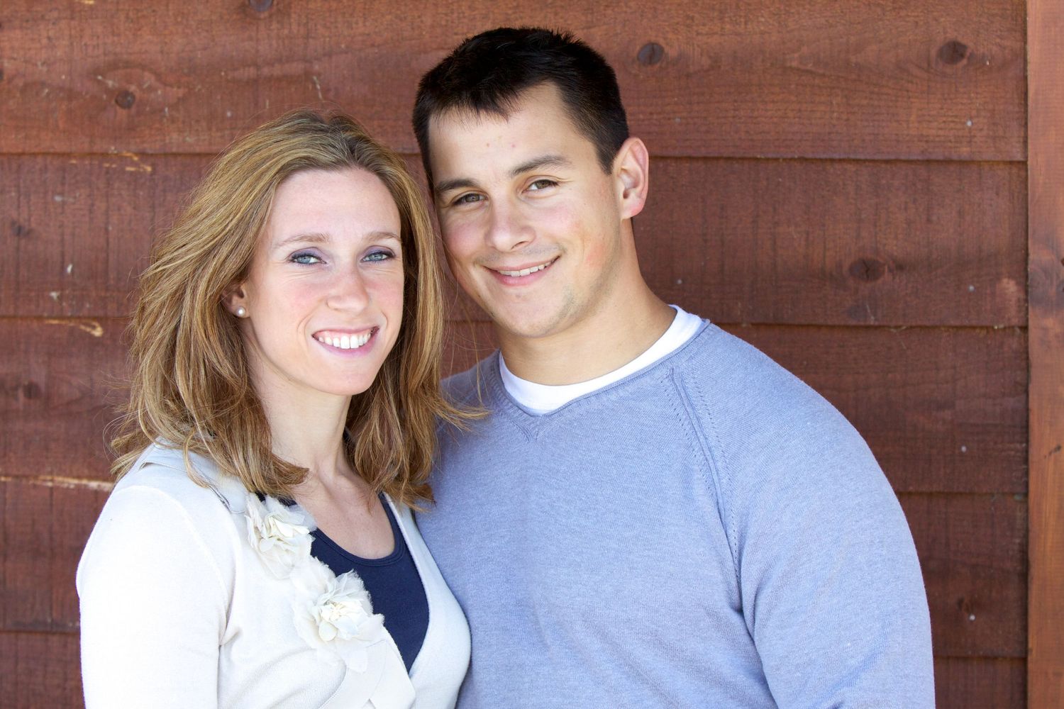 Couple in casual attire posing against rustic wooden wall background.