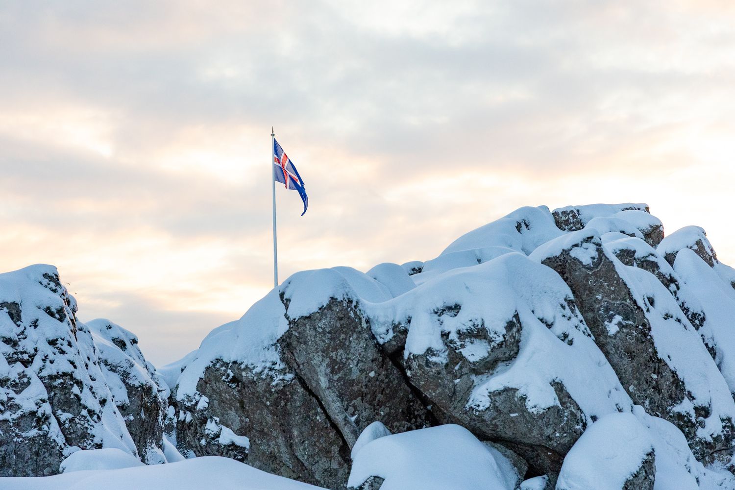 A flag waves atop snow-covered rocky peaks against a cloudy sunset sky.