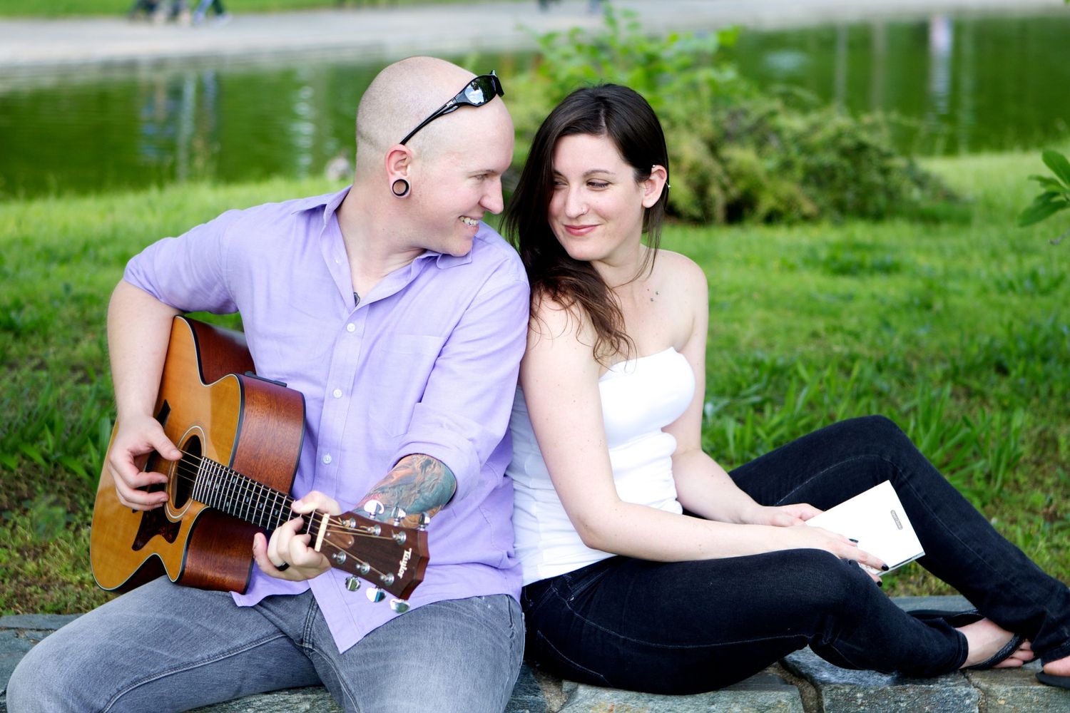 A couple sits on grass while one plays acoustic guitar in a romantic outdoor setting.