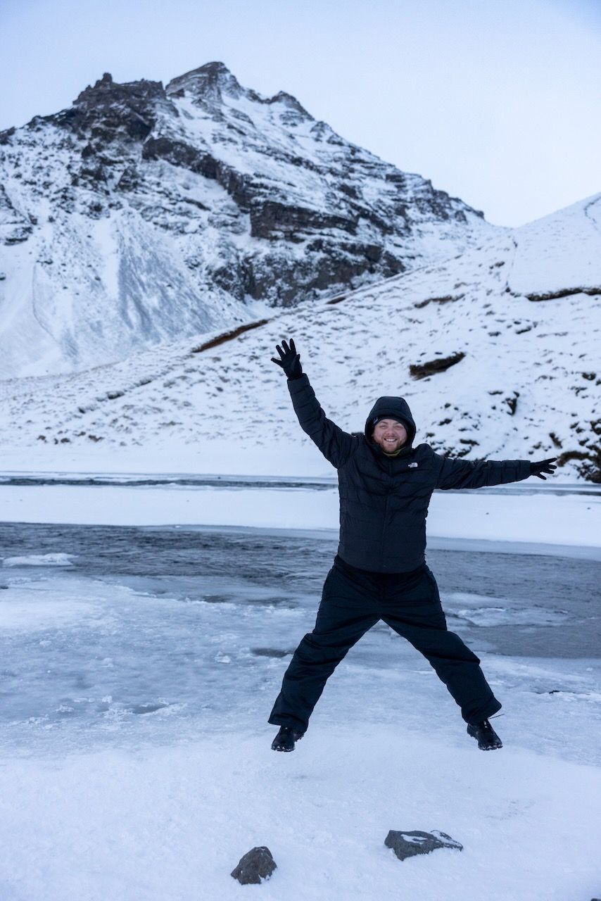 Person making snow angels and playful poses on frozen ground with snowy mountains in background.