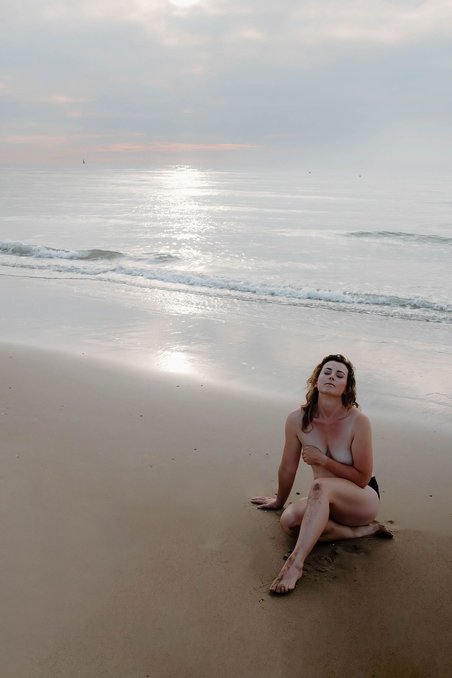 A woman sits on a sandy beach, gazing out at the ocean with the sun reflecting on the water.