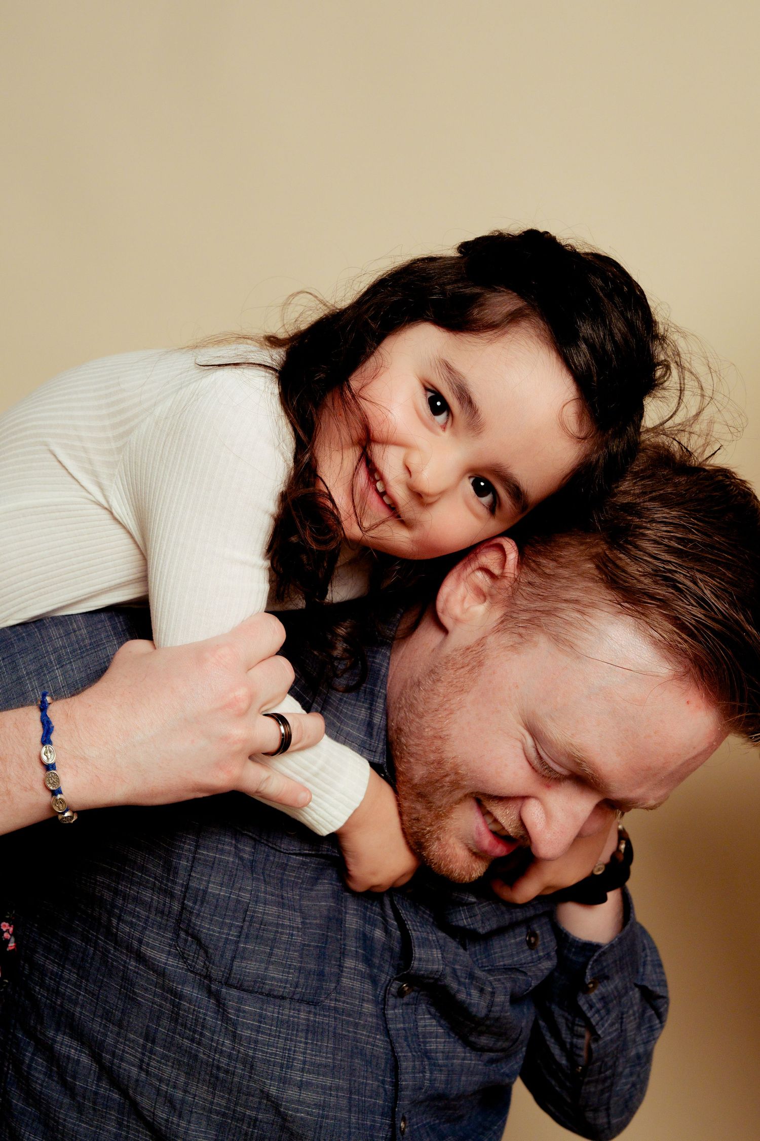 A smiling young girl with dark curly hair rides piggyback on a man's shoulders, both laughing together indoors.