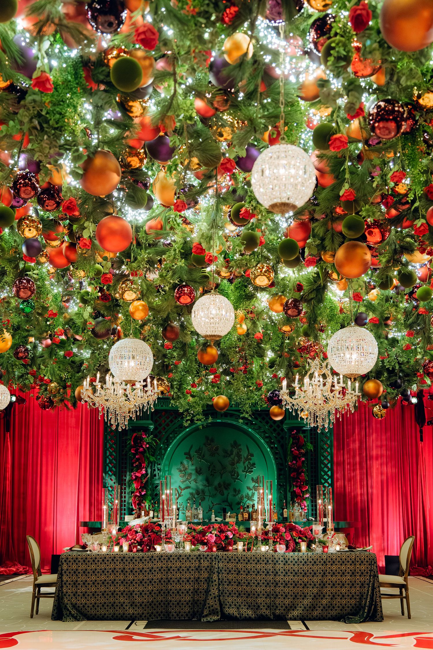 Opulent event space with crystal chandeliers, orange ornaments, and red drapery under garden-inspired ceiling.