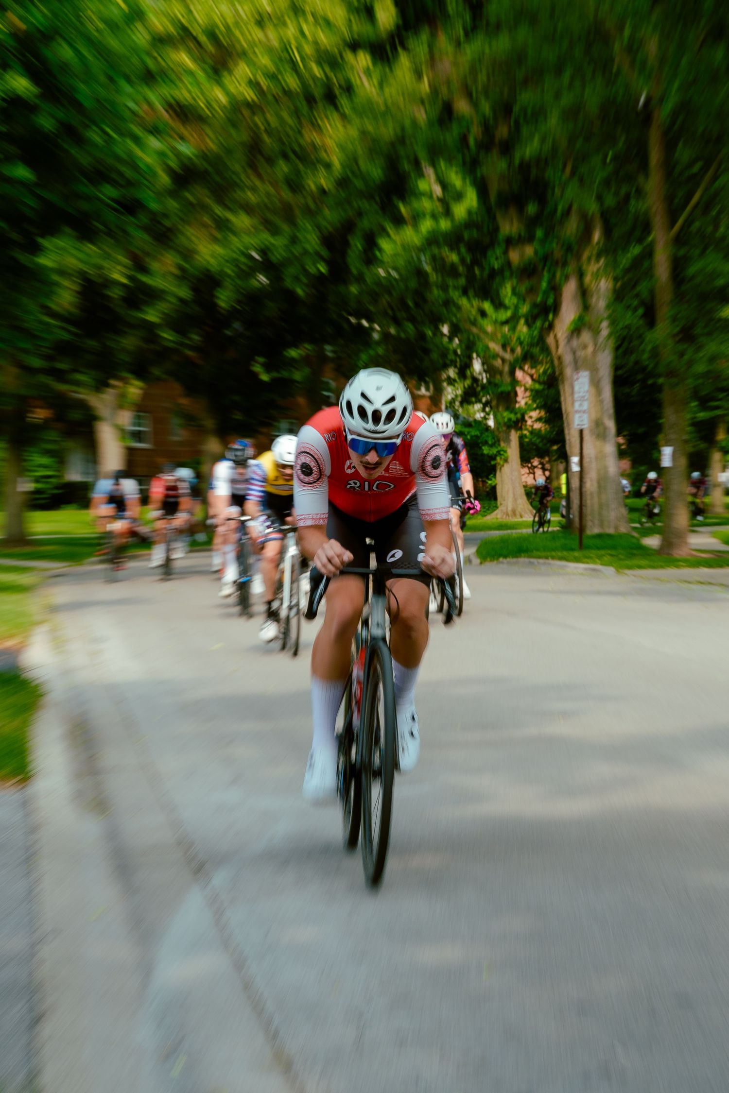 Cyclist in racing gear leads a pack on a tree-lined road with motion blur effect.