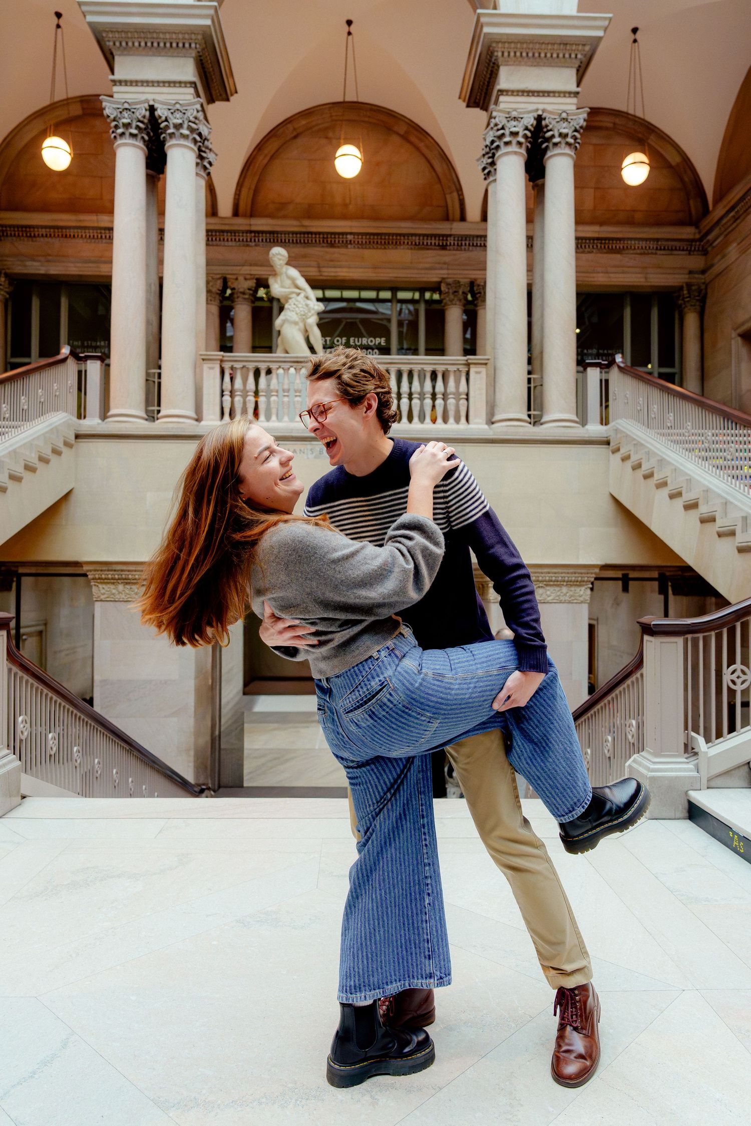 A couple poses playfully in a grand historic building interior with ornate columns and arches.