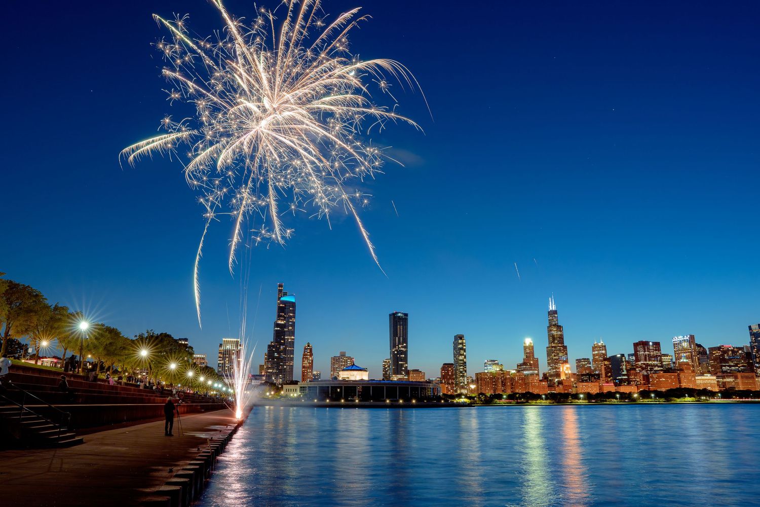 Fireworks illuminate the Chicago skyline at night along Lake Michigan with bright bursts reflecting on the water.