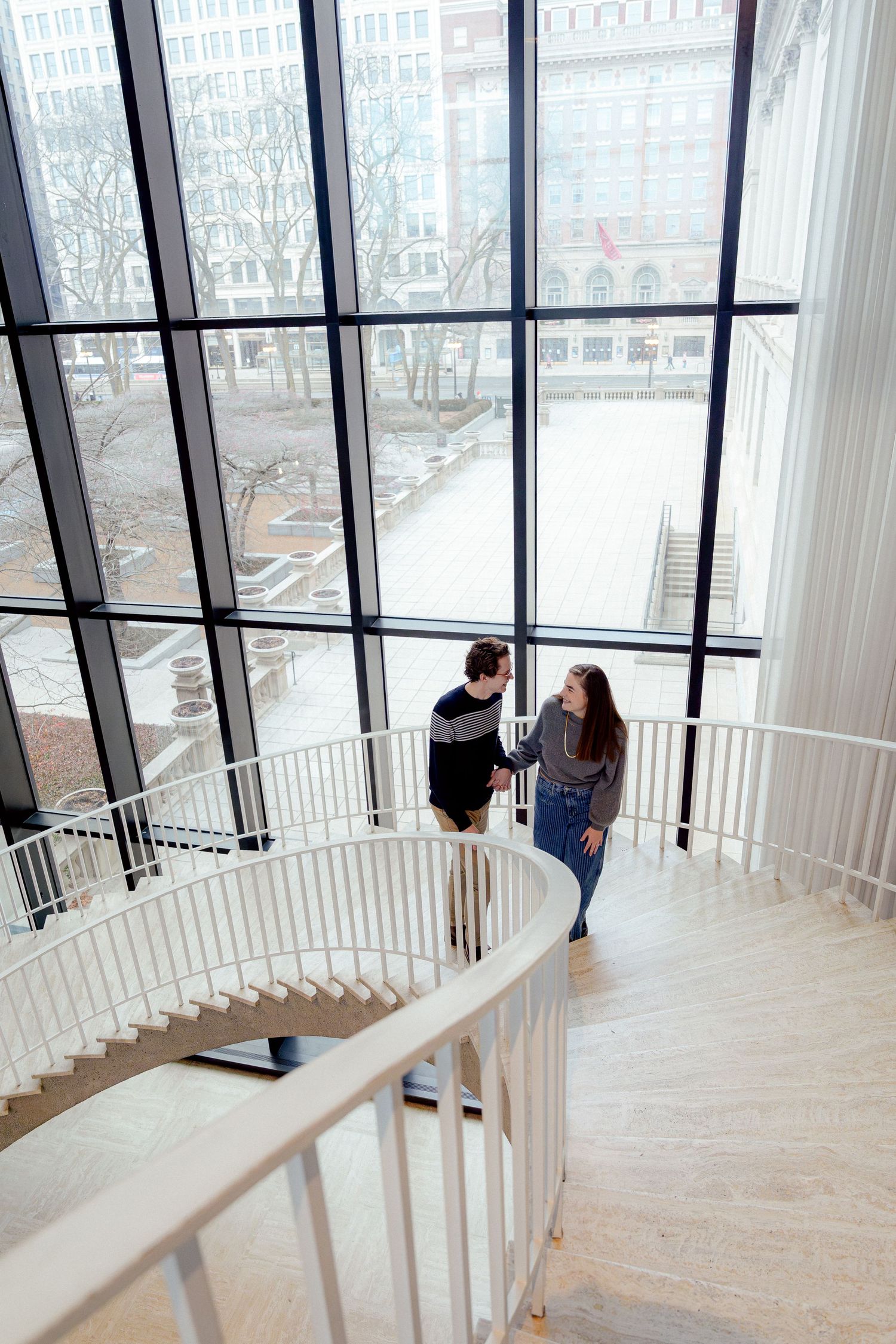 A couple stands on an elegant spiral staircase with large windows overlooking the city.