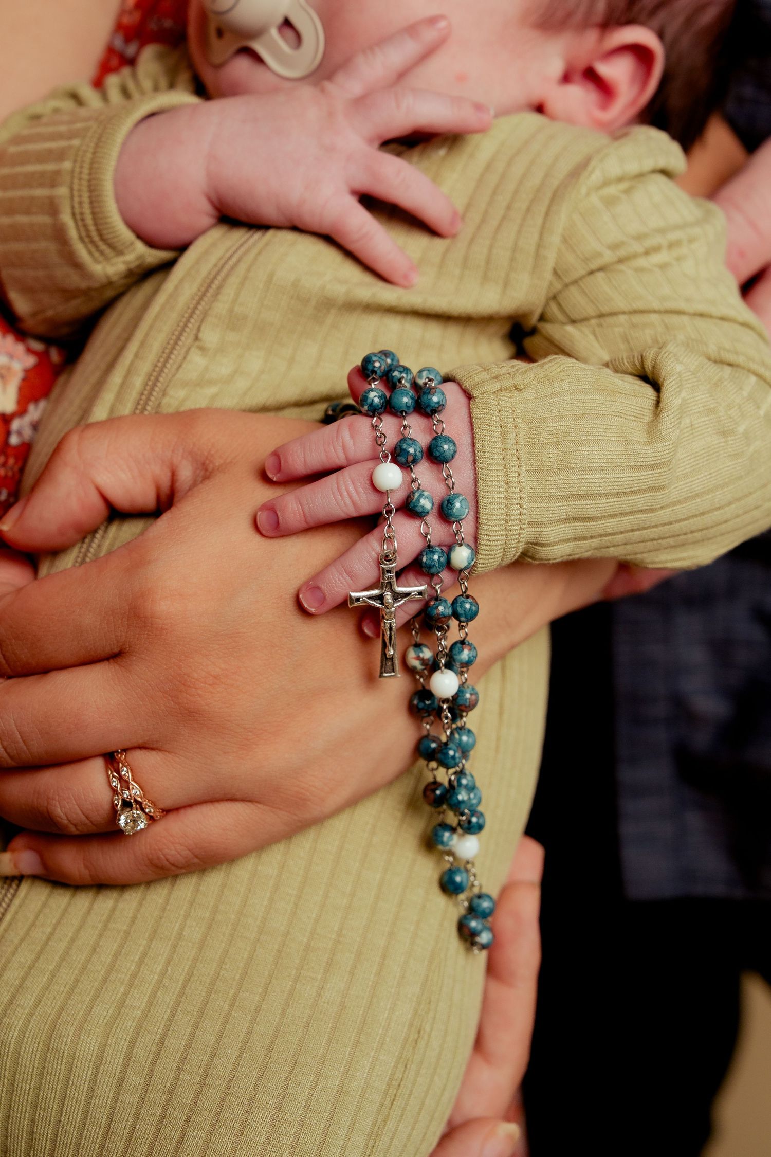 Adult hands holding a newborn baby with a blue rosary beaded necklace with silver crucifix cross.