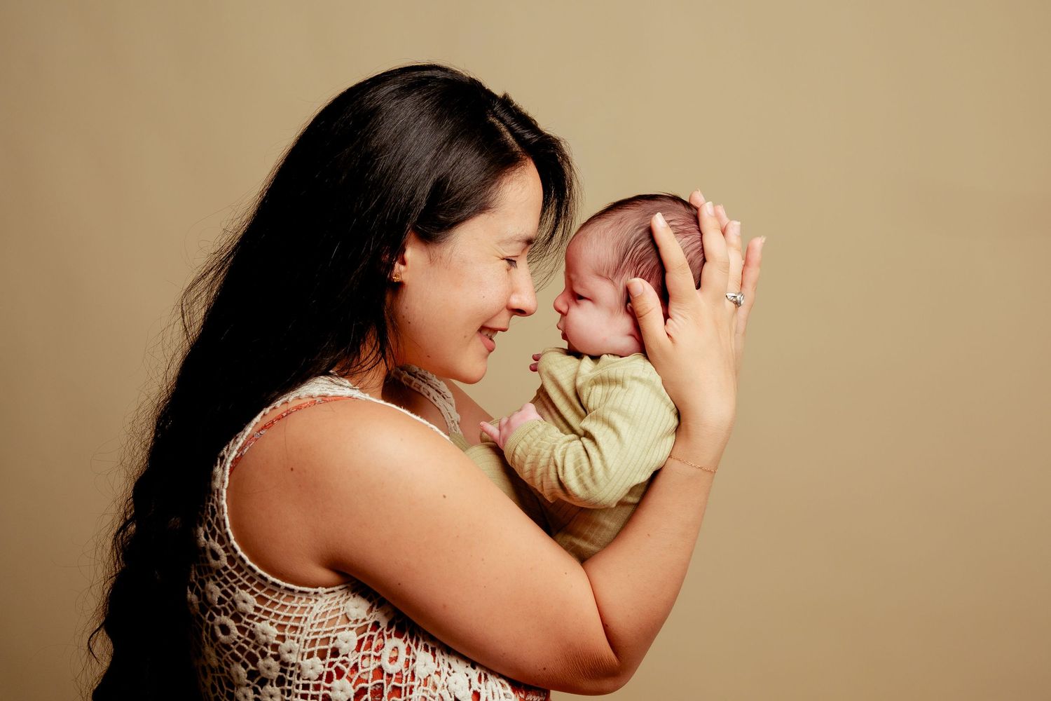 A smiling mother tenderly holds her sleeping newborn baby close to her face against a warm beige background.