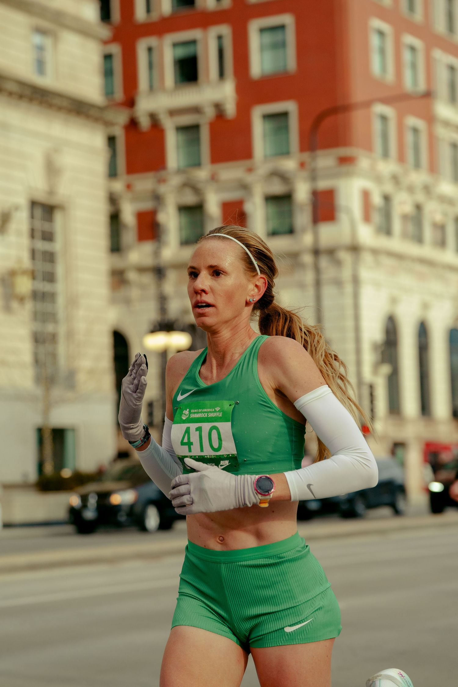 Female marathon runner in green outfit and bib number 410 races through city streets past red brick building.